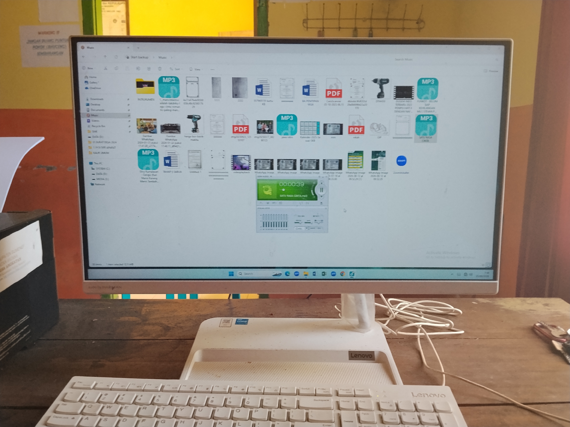The image shows a computer monitor displaying a file explorer window, likely on a desk in Laok Jangjang, Indonesia. The monitor, a white Lenovo All-in-One PC, is positioned in front of a wooden desk. The screen shows a folder named "Music" with various files including MP3 audio files, PDF documents, and image files. Several icons for different applications, including a web browser, are visible in the taskbar at the bottom of the screen. A white Lenovo keyboard is placed in front of the monitor. A black speaker is to the left of the monitor. Cables are visible on the desk, some connected to the computer. The background consists of a wall with some posters and yellow and green painted sections. The lighting suggests an indoor environment, possibly a room in a home or office. There are no people visible in the image.