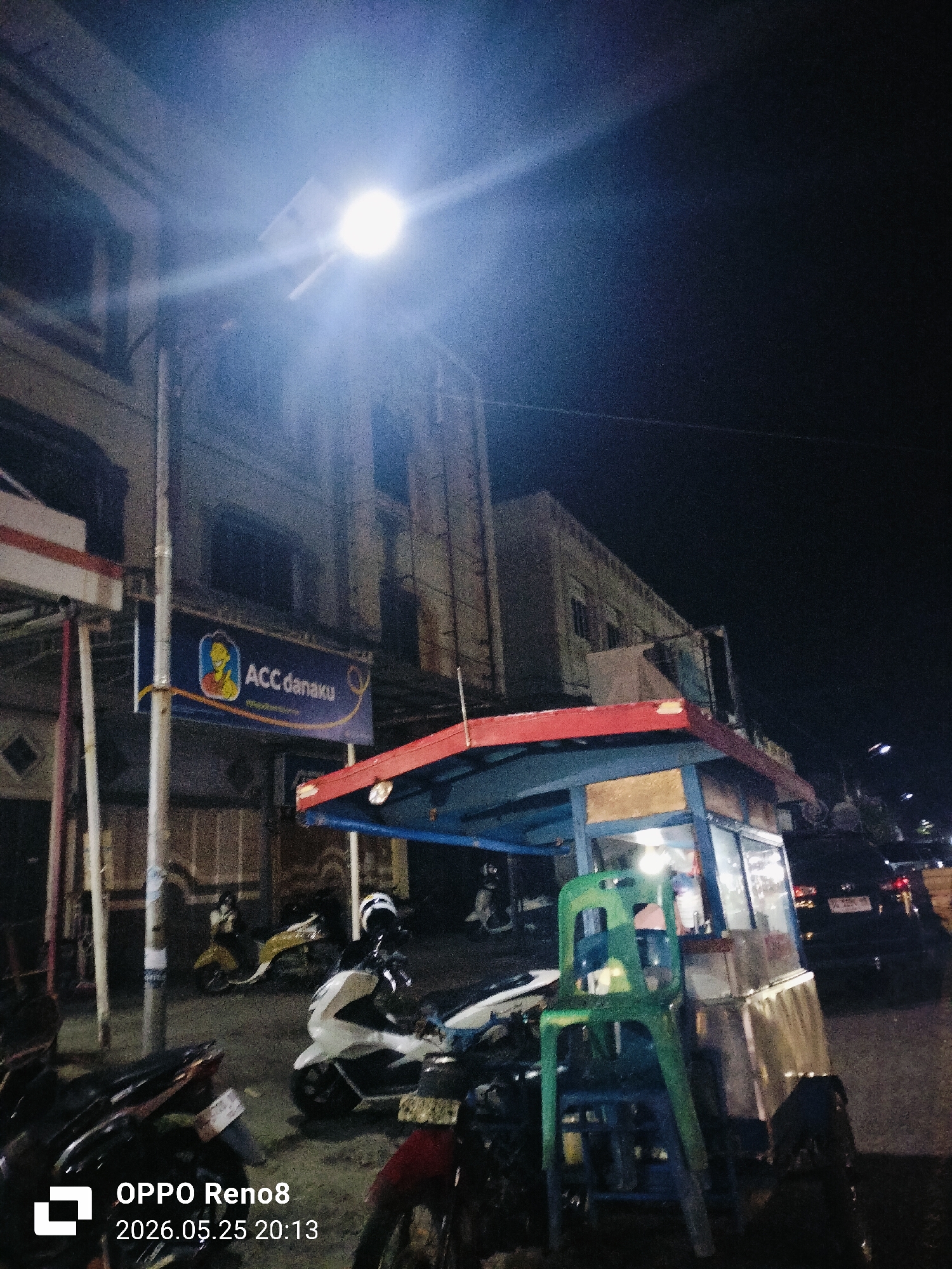 The image shows a street scene in Lhokseumawe, Indonesia at night. A bright street light illuminates a food vendor's cart with a red and blue awning. A green plastic chair is stacked beside the cart. Several motorcycles are parked nearby. A building with a sign reading "ACC danaku" is in the background. A car is visible on the street. The image includes a timestamp "OPPO Reno8 2026.05.25 20:13".