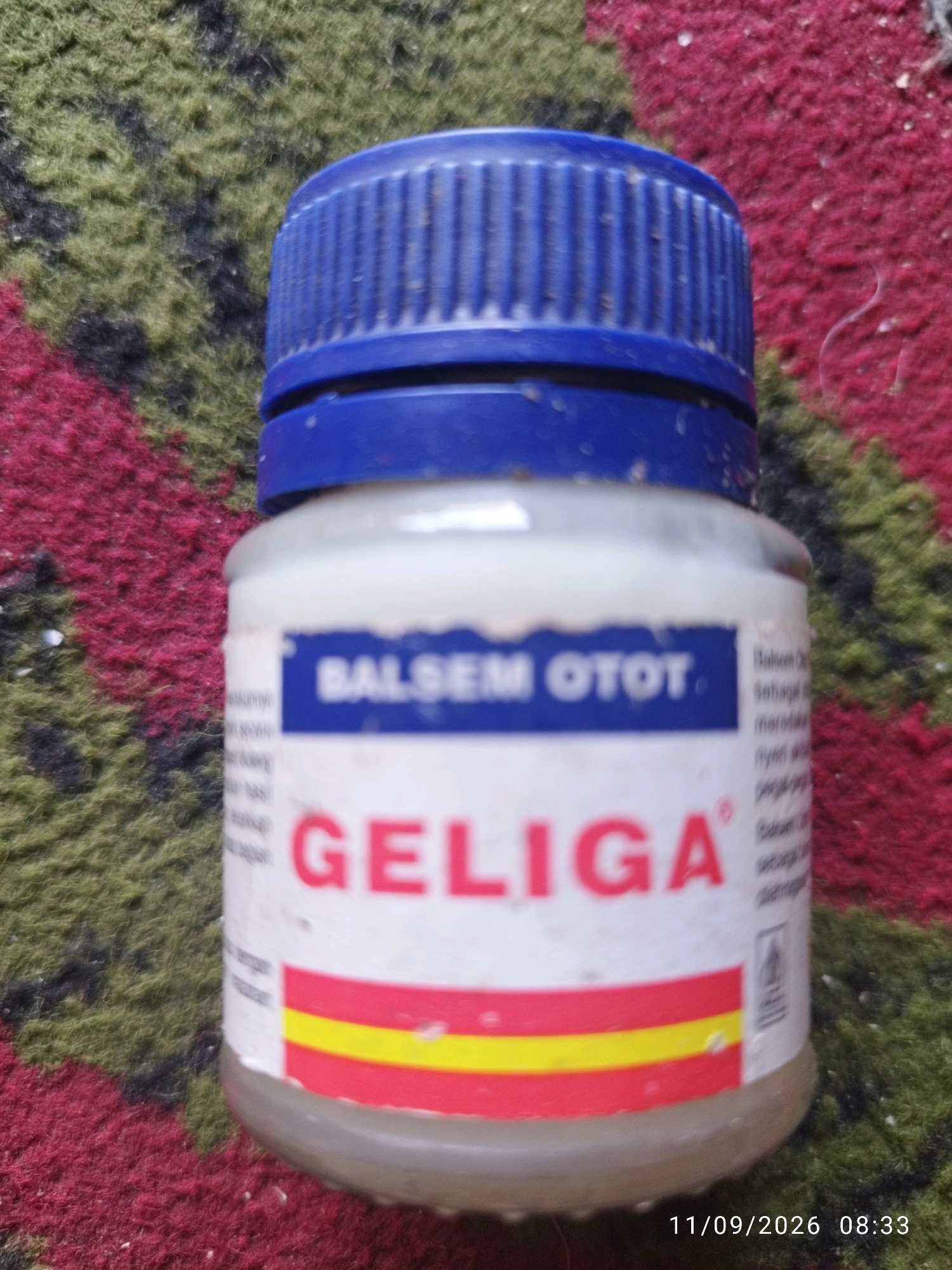 A close-up, high-angle shot shows a bottle of muscle balm, identified as "GELIGA BALSEM OTOT," resting on a textured, multi-colored surface. The bottle has a blue plastic screw-top lid and a white label with red and yellow stripes. Text on the label, partially obscured, includes "GELIGA" in large red letters and "BALSEM OTOT" in smaller blue letters. The background is a carpet or rug with a pattern of green, black, and red fibers. The bottom right corner of the image displays a timestamp: "11/09/2026 08:33". There is no discernible indication of the city of Mojokerto, Indonesia, within the visual elements of the image itself.