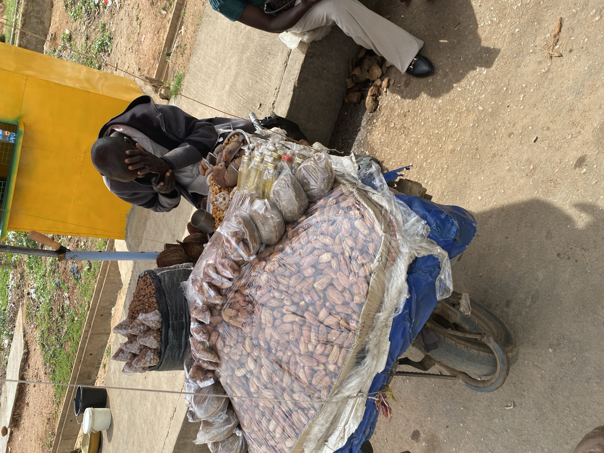 The image depicts an outdoor market or roadside vending scene in Bukuru, Nigeria, under bright daylight. A vendor's stall, likely a modified bicycle or tricycle, is laden with various goods for sale. The dominant item is a large, clear plastic-wrapped bag filled with numerous brown, elongated fruits, appearing to be dates. Alongside these, smaller clear plastic bags contain what look like peanuts or other small nuts, and several glass bottles filled with a yellow liquid are arranged. Coconut shells are also visible among the merchandise.
A man, presumably the vendor, is seated on the ground to the left of the stall. He wears a dark jacket over a striped shirt, with his head down and hands covering his face, suggesting rest, contemplation, or possibly distress. Partially visible to the right is another person sitting on the ground, wearing a green top and white pants.
The ground is a light-colored, rough surface, possibly concrete or compacted earth. Strong shadows indicate a sunny day. In the background, there's an unpaved area with sparse green vegetation and some debris, next to a concrete drainage ditch. A yellow wall or structure is visible to the far left. Further down the path, two plastic buckets, one black and one white, are placed. No discernible text is visible on any items or signs.