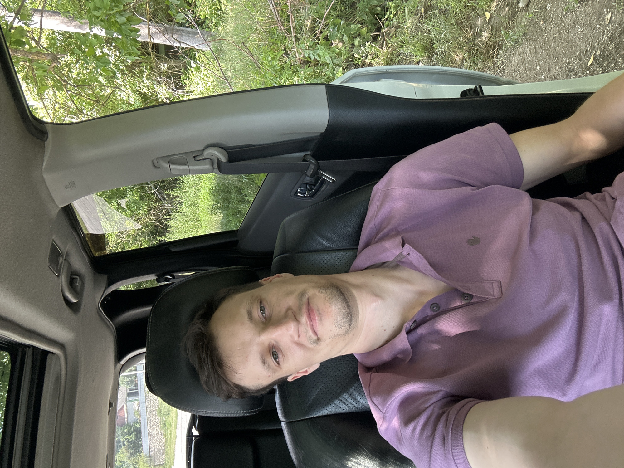A man with short brown hair and light skin is sitting in the driver's seat of a car. He is wearing a purple polo shirt. He is looking at the camera and smiling slightly. The car is parked on a dirt road, with trees and greenery visible on either side. A house is visible in the background on the left. The scene appears to be in Makiv, Ukraine.
