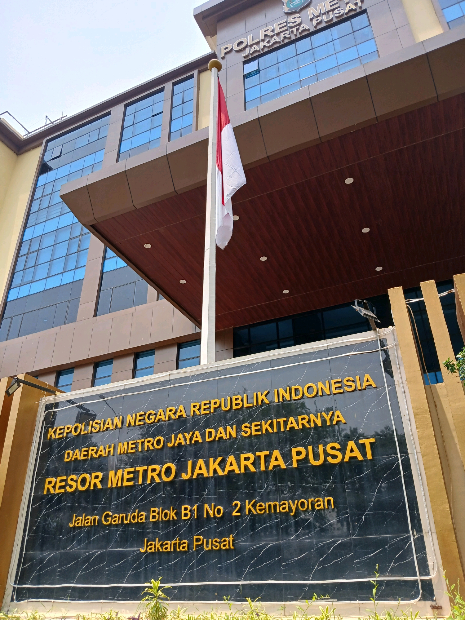 The image shows the exterior of the POLRES METRO JAKARTA PUSAT building, located in the city of Central Jakarta, Indonesia. The building is a modern, multi-story structure with a facade of blue tinted glass and brown cladding. A flagpole with the Indonesian flag (red and white) stands prominently in front of the building. Below the building, a black marble sign with raised yellow lettering displays the official name of the police station: "KEPOLISIAN NEGARA REPUBLIK INDONESIA DAERAH METRO JAYA DAN SEKITARNYA RESOR METRO JAKARTA PUSAT" and the address "Jalan Garuda Blok B1 No 2 Kemayoran Jakarta Pusat". The overall scene depicts an official government building under a bright, overcast sky. There are no people visible in the image.
