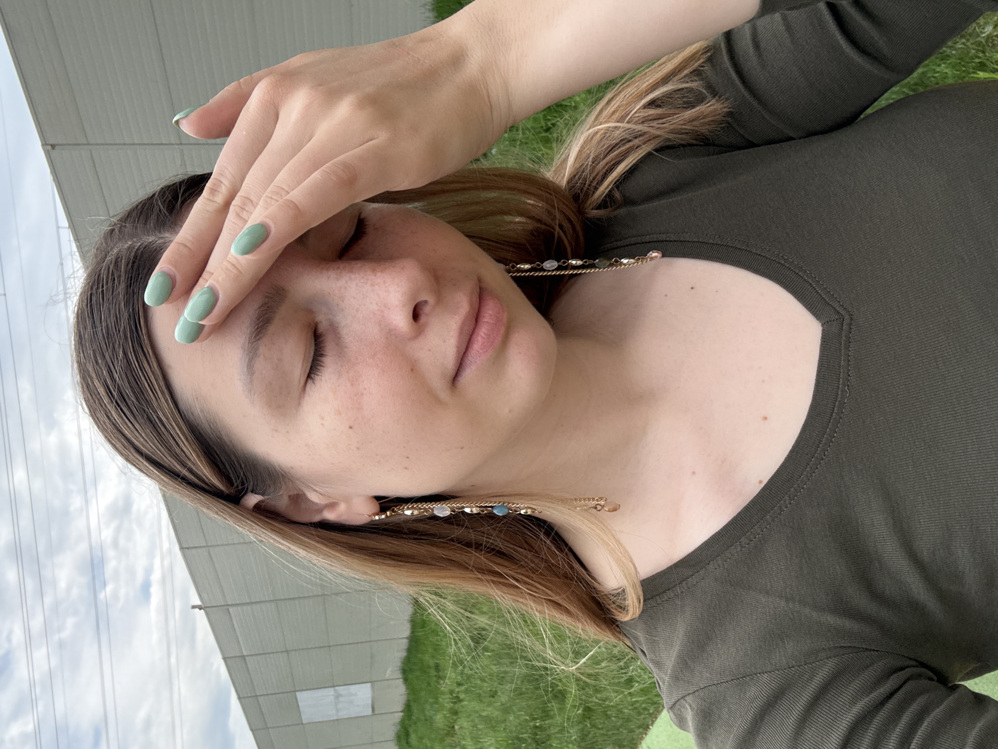 A young woman is outdoors, likely in a park or grassy area. She wears a long-sleeved, dark green top and has a gold-toned hair clip with gemstones or beads visible near her ear. Her left hand is raised to her forehead, possibly shading her eyes. Her fingernails are painted a light green color. The background includes a building with white siding and patches of green grass. The location in Moscow, Russia cannot be determined from this image alone.