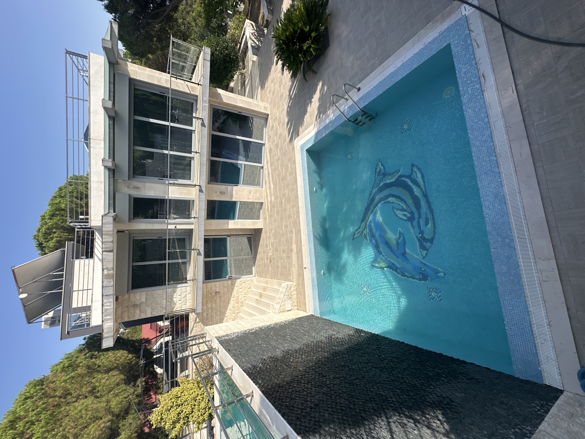 This is a medium-angle, eye-level, long shot of a modern villa with a swimming pool in Brca, Montenegro. The villa is a multi-story building with a light stone facade, large glass windows, and multiple balconies with metal railings. A flat roof with solar panels and air conditioning units is visible. The swimming pool is in the foreground, filled with turquoise water and features a mosaic of two blue dolphins at the bottom. The pool is surrounded by light-colored paving stones. To the left of the pool, a dark stone wall slopes upwards, leading to a retaining wall with a metal railing and a small tree. Stairs made of light stone connect the pool area to a higher level. Lush green trees are visible behind the villa and to the sides. The sky is clear and blue. No people are present in the scene.