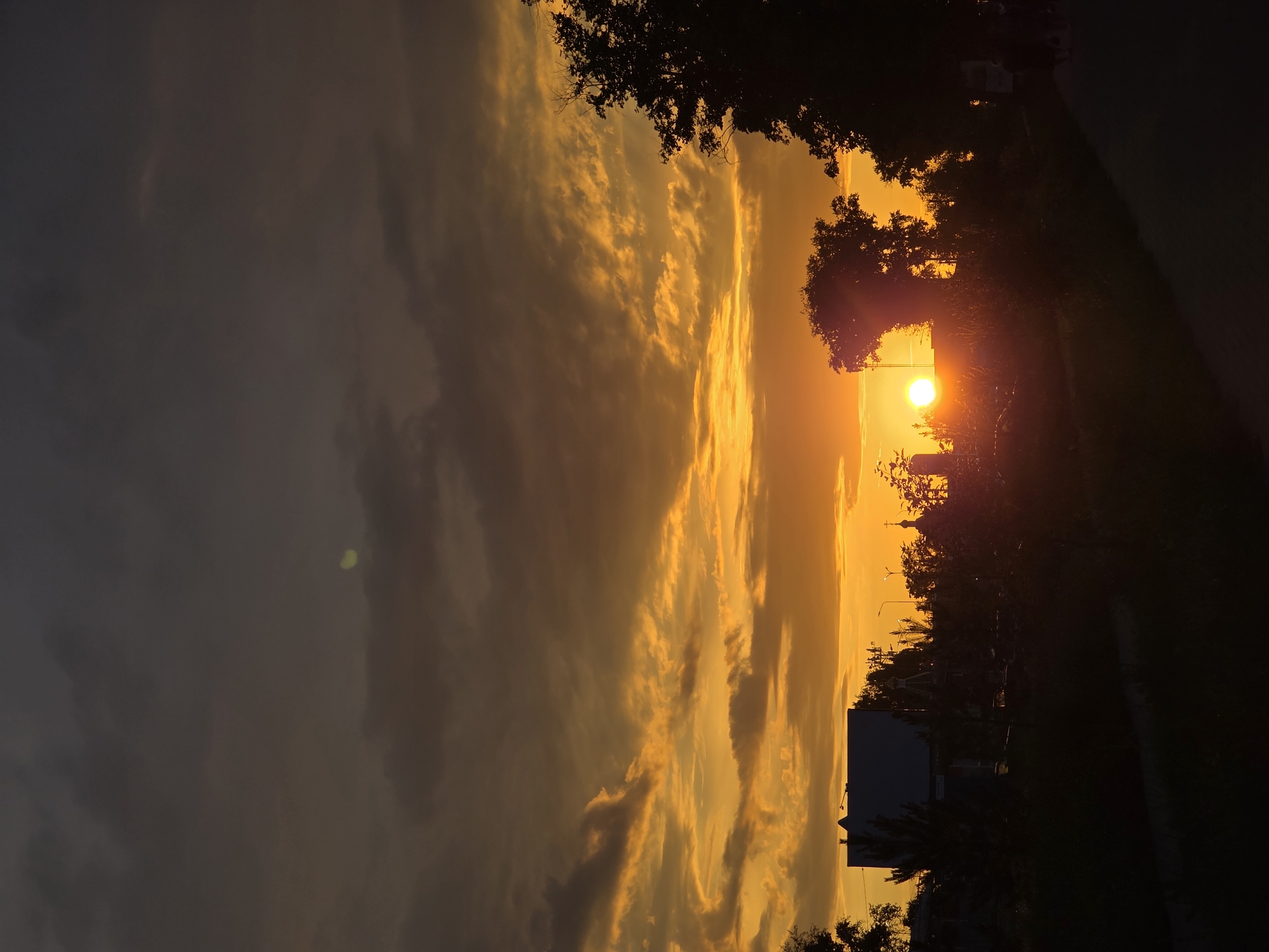 The image captures a sunset over a residential area in Chaikovsky, Russia. The sky is filled with dramatic clouds painted in shades of orange, yellow, and dark gray. In the foreground, silhouetted trees and buildings obscure the details of the urban landscape. A bright, round sun is visible near the horizon, casting a warm glow. The foreground also shows a dark, paved path, suggesting a pedestrian walkway. The overall scene is one of a fading day, with the silhouetted structures and vegetation creating a stark contrast against the vibrant sky.