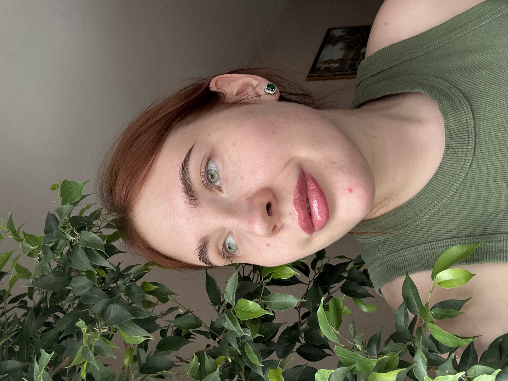 A woman is posing in front of a leafy green plant. She has fair skin, auburn hair, and green eyes. She is wearing a green tank top and a green gemstone earring. The background appears to be an interior setting, possibly a room in a house or apartment. A framed image can be seen in the background near her ear. The image does not depict the location in the city of Chaikovsky, Russia.