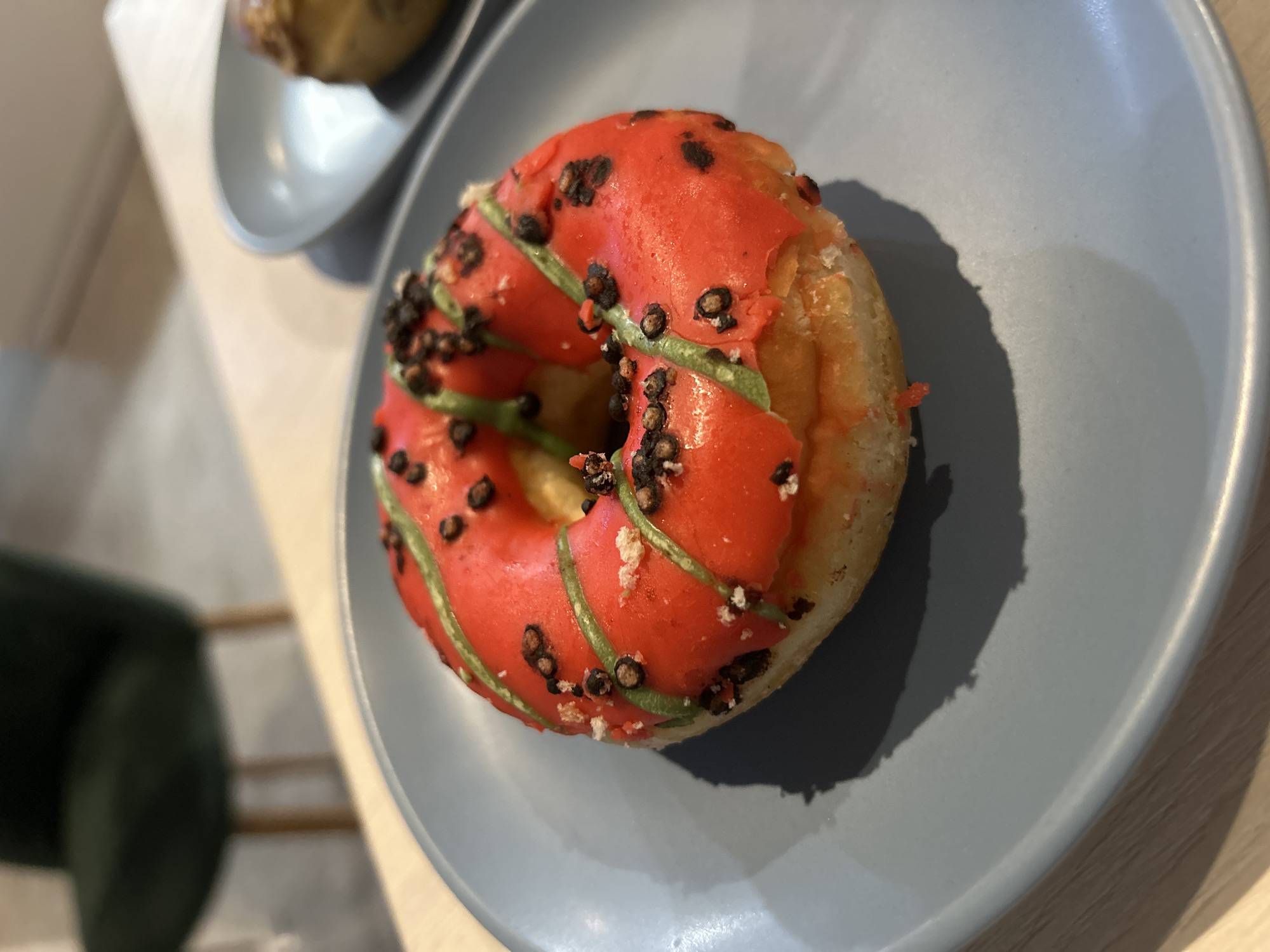 A close-up shot captures a single, iced doughnut resting on a light blue plate. The doughnut is topped with bright red icing, adorned with thin green stripes and dark, granular sprinkles, possibly chocolate or coffee grounds, and some white crumbs. The doughnut has a visible hole in the center and a slightly broken edge. The plate casts a soft shadow on the light-colored wooden table. In the blurred background, a dark green chair with wooden legs is visible, suggesting an indoor setting, possibly a cafe or restaurant. The city of Chaikovsky, Russia, is not discernible from the image.