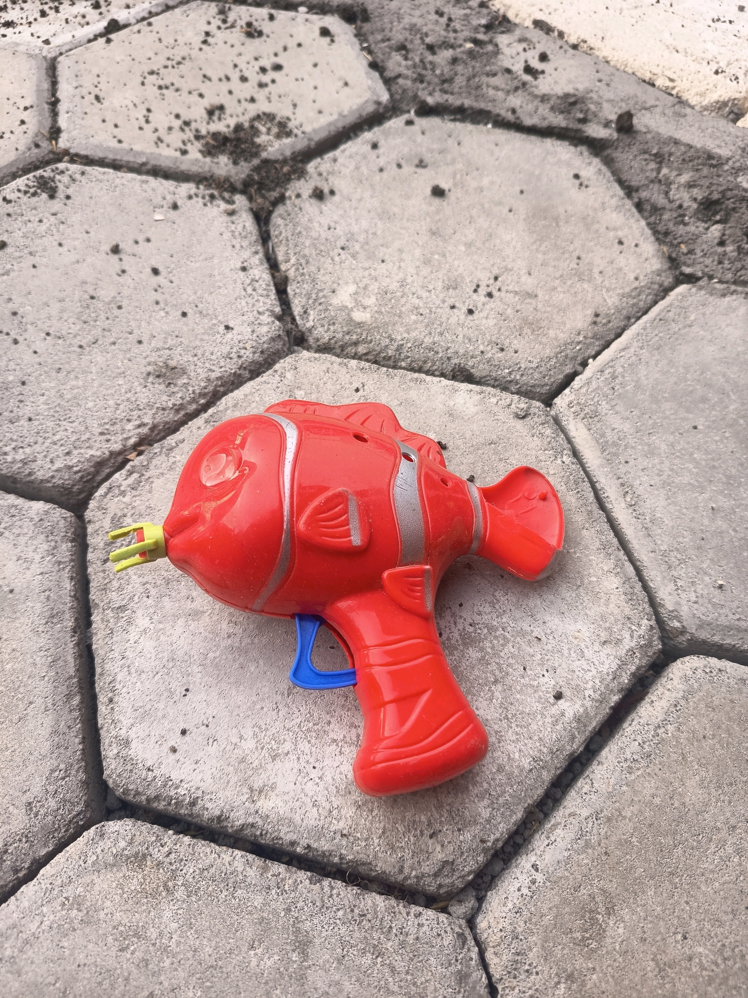A red toy water gun shaped like a clownfish lies on a patio made of interlocking hexagonal concrete pavers. The pavers are worn and speckled with dirt and debris. The water gun is bright red with silver accents on its fins and body, and a blue trigger. The front of the water gun has a yellow nozzle.
