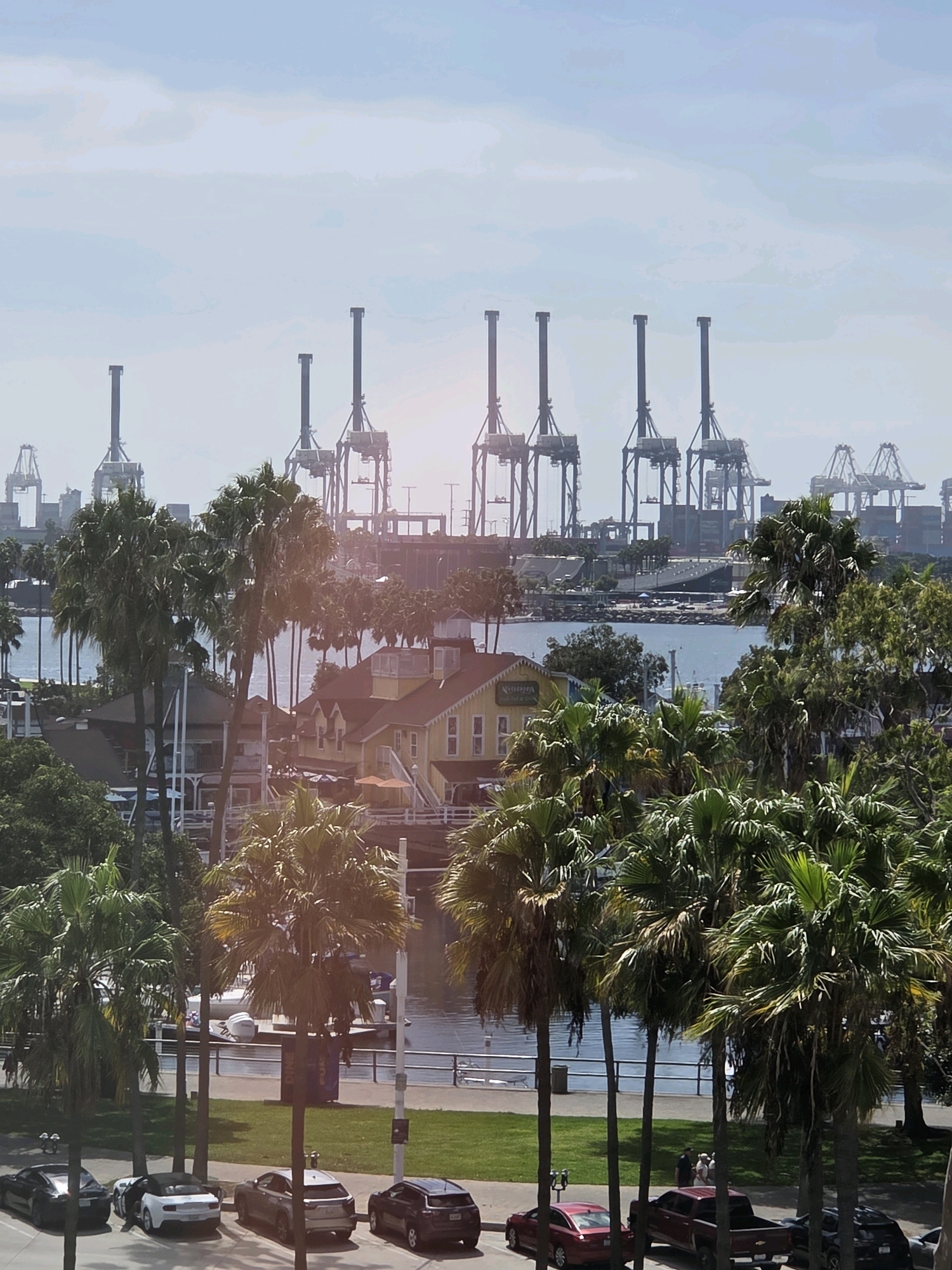 This image captures a wide view of the Port of Long Beach, California, USA, under a hazy sky. In the foreground, a line of palm trees frames the scene. Beyond the trees, several buildings with red roofs and yellow walls are visible, suggesting a waterfront commercial area. A body of water, likely the harbor, separates these buildings from the main port activity. Towering over the water are multiple large shipping cranes, characteristic of a busy port. Several boats are docked in the water near the buildings. In the lower part of the image, a street runs alongside the harbor, with several cars parked along it, including a black sedan, a white convertible, a silver sedan, and a dark SUV. A red sedan and a red pickup truck are parked further down the street. A grassy area with a fence separates the street from the water. In the background, more cranes and industrial structures are visible, indicating the vastness of the port.