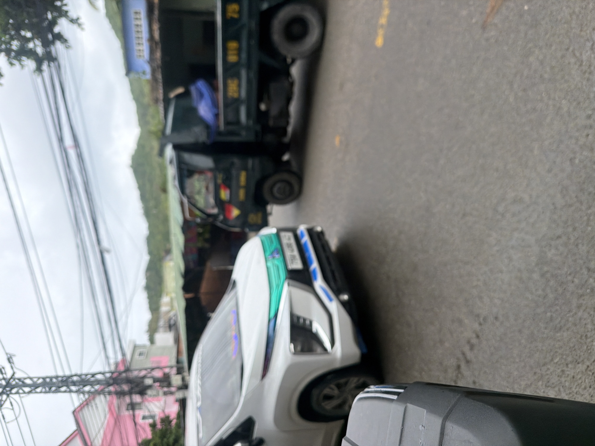 In Nha Trang, Vietnam, a blurry street scene depicts a white Mitsubishi Xpander parked in the foreground, facing right. The car features a turquoise accent on its grille and blue accents on its front bumper. Behind it, a dark-colored utility truck with yellow lettering on its side, possibly a license plate, is parked. The truck's bed contains a blue tarp. In the background, a pink building stands next to a utility pole with numerous wires. Further in the distance, green, tree-covered hills are visible under an overcast sky. The road surface is asphalt with faint yellow markings.