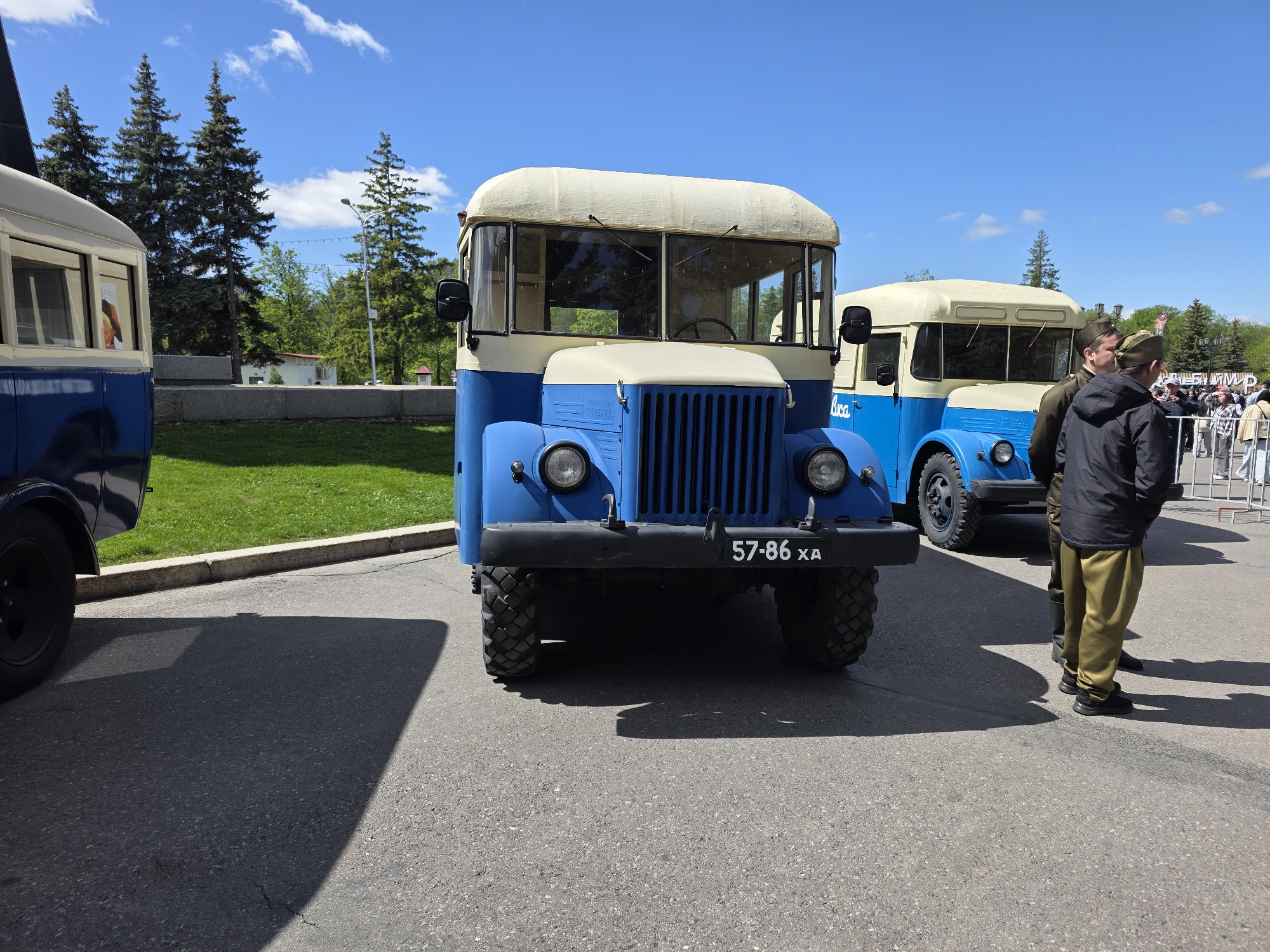 The image shows two vintage blue and white buses parked on an asphalt surface. The first bus is in the foreground, facing the camera. It has a license plate reading "57-86 XA." A second bus, similar in design, is visible in the background to the right of the first bus. Two men wearing olive green military-style uniforms are standing near the second bus. A partially obscured bus is on the left side of the picture. Behind the buses, there is a green lawn with a concrete border. Tall pine trees are visible behind the lawn, and a blue sky with white clouds is overhead. The city of Ufa, Russia, is implied as the buses are often used in parades and celebrations there.