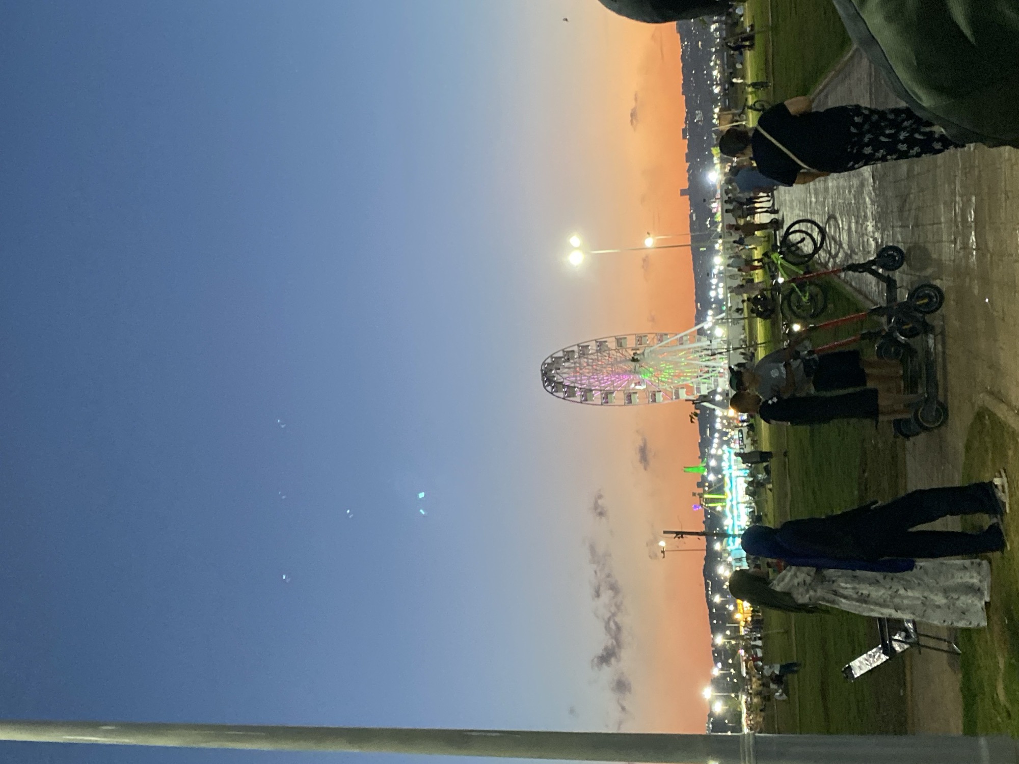 A wide shot captures a twilight scene in the Hussein Dey, Algeria area, with a large Ferris wheel illuminated with colorful lights dominating the background. The sky transitions from a deep blue at the top to a vibrant orange and pink near the horizon. In the foreground, several people are visible, including a woman in a patterned dress and headscarf standing next to another woman in a dark hijab. Two electric scooters are parked on the paved walkway. The ground is a mix of grass and the paved path. Further back, a crowd of people are scattered across the grassy area and near a row of illuminated structures, possibly market stalls or amusement park rides. Streetlights are also visible, adding to the illuminated atmosphere. The overall impression is of a lively public space during an evening event.