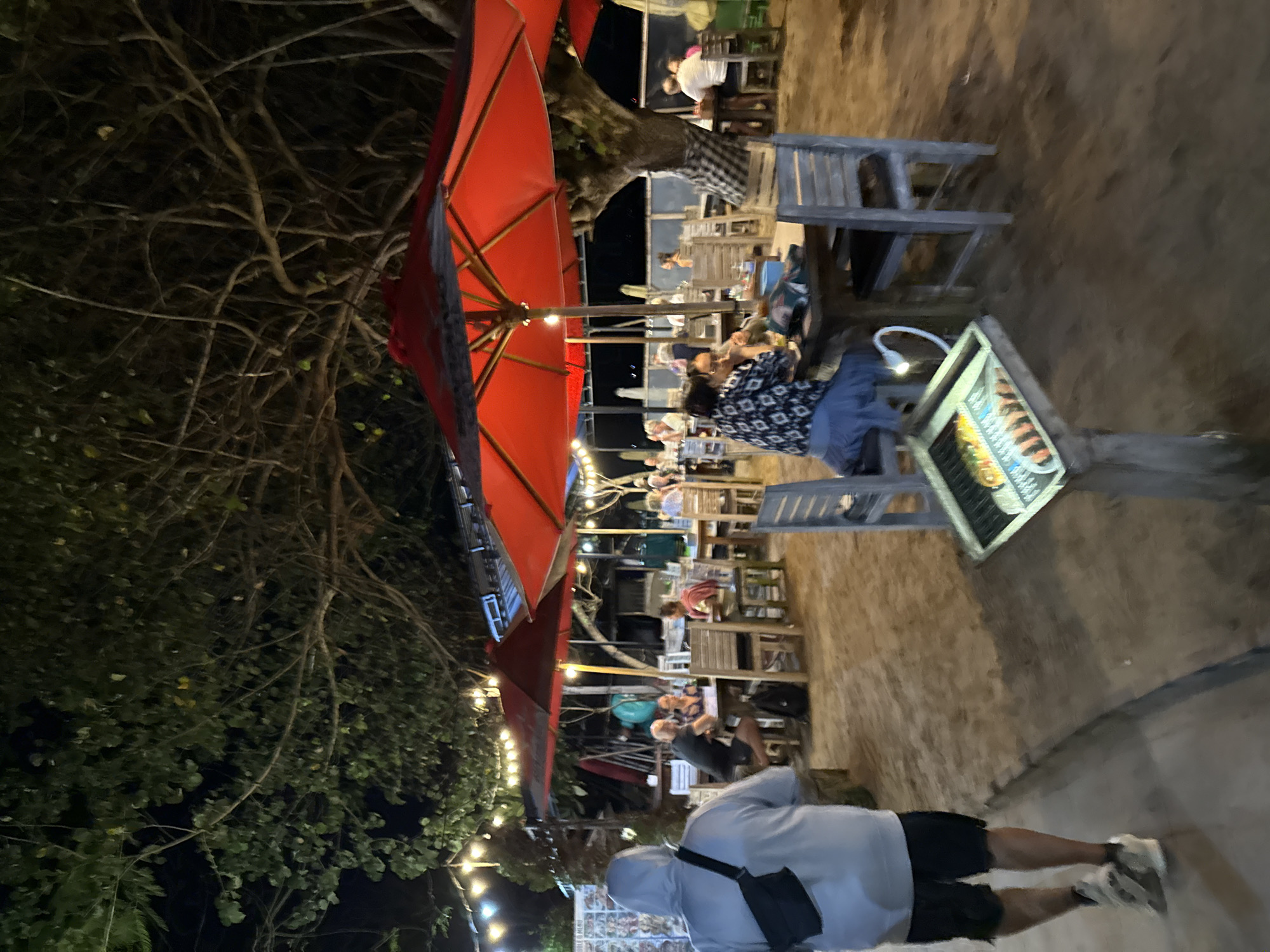 An outdoor restaurant at night in Denpasar, Indonesia, features a sandy floor and thatched-roof structures, illuminated by strings of light bulbs and red umbrellas providing shade over dining tables. Several patrons are seated at tables, some engaged in conversation. A person wearing a grey hooded sweatshirt and black shorts walks away from the camera, while another person in a black and white patterned shirt and blue skirt is seated at a table. A menu display is positioned on a wooden stand.