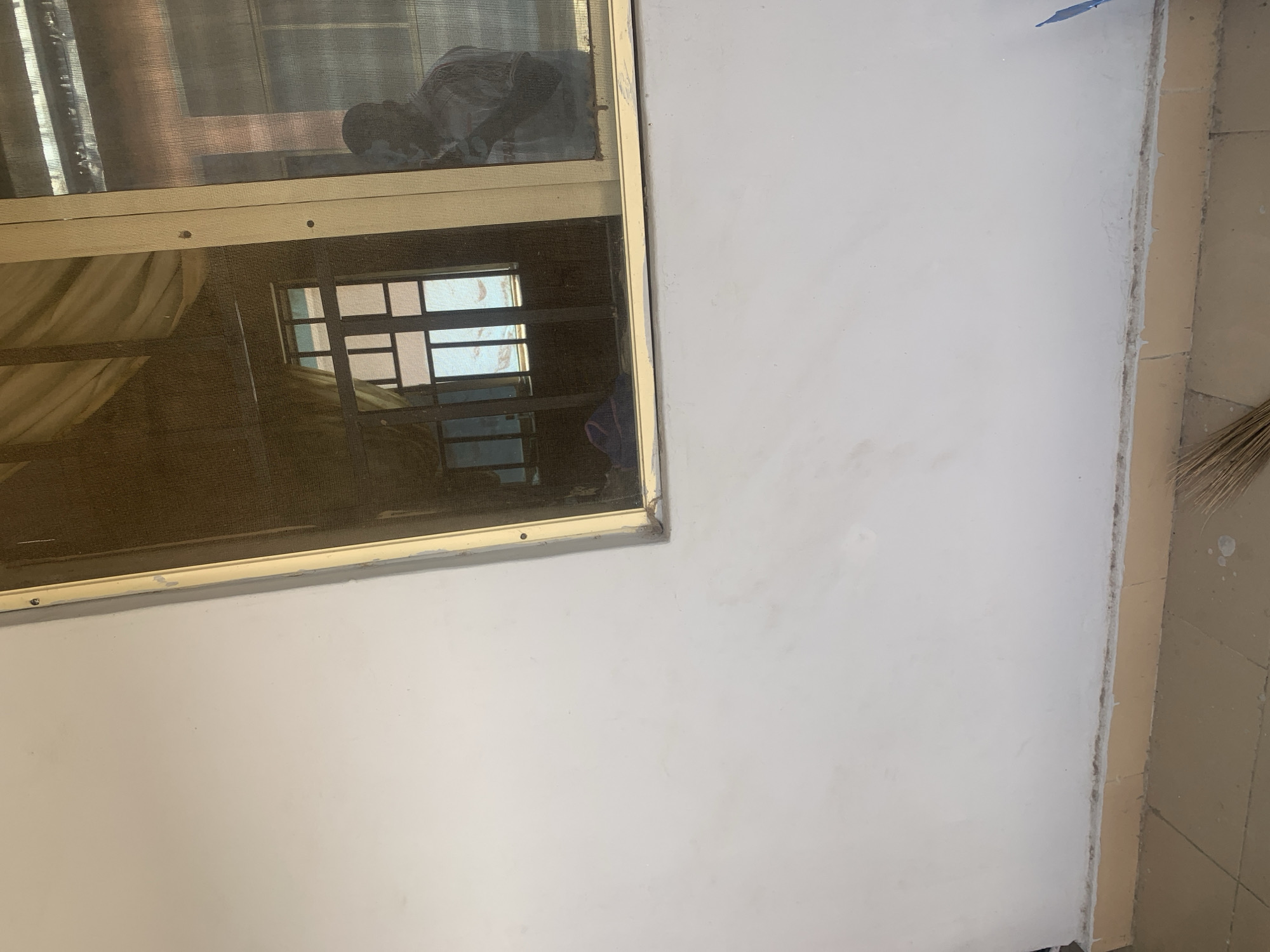 The image shows a close-up, eye-level view of a white wall adjacent to a screened window. The window has a light-colored frame and is slightly dirty. Reflections in the screen show an interior scene with a patterned curtain or drapery, a window with multiple panes, and a dark silhouette of a person. The bottom of the wall meets a beige tiled baseboard and floor, with a broom or similar cleaning tool resting on the floor near the right edge. The overall scene suggests an outdoor view of a building in Jauro Yinu, Nigeria, likely a residential or commercial space.