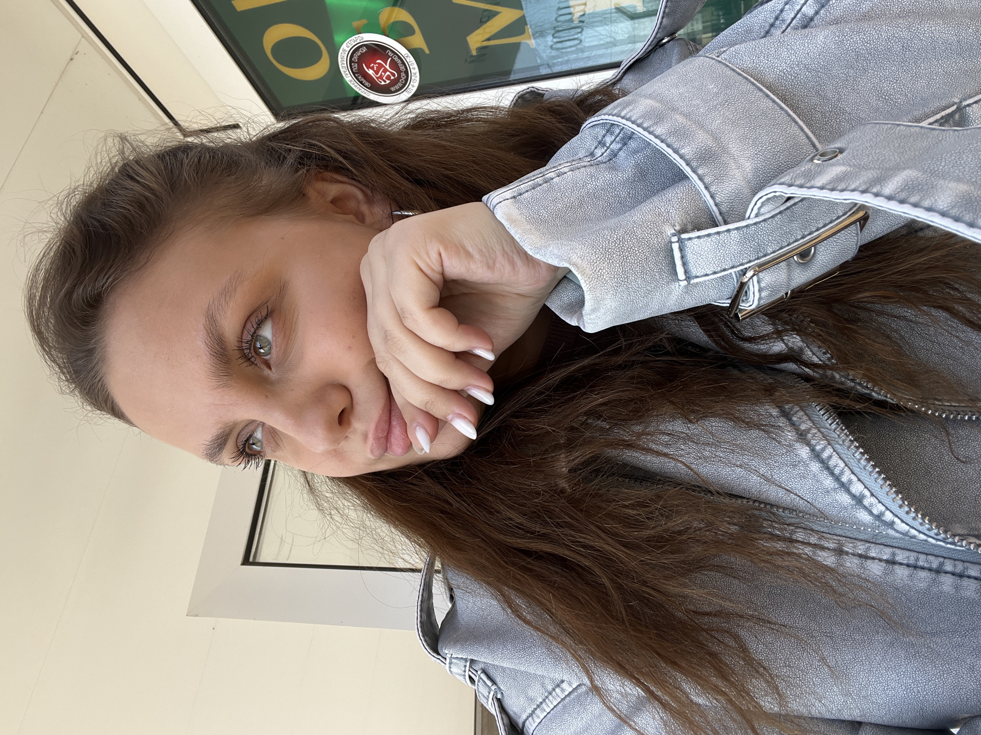 The image is a selfie of a young woman wearing a silver metallic jacket. She has long brown hair and light-colored eyes. Her right hand is gently resting against her cheek. Behind her, there is a window with some lettering and a circular sticker with the inscription: "ПРЕДПРИЯТИЕ «ΓΕΠΑΡΩ» / ЕКТ ПОД ОХРАНО / ОХРАННОЕ / ОБЬЕКТ / GEPARD RU / 4-51-30 / ЕЛ 4-15-24". The text "N" and "10:00" are also visible on the sticker. Based on the Cyrillic text and "GEPARD RU", the location is likely in Russia. The Gepard company is a company for security services and according to the contact information located on the sticker, the company is located in the city of Chaikovsky, Russia.