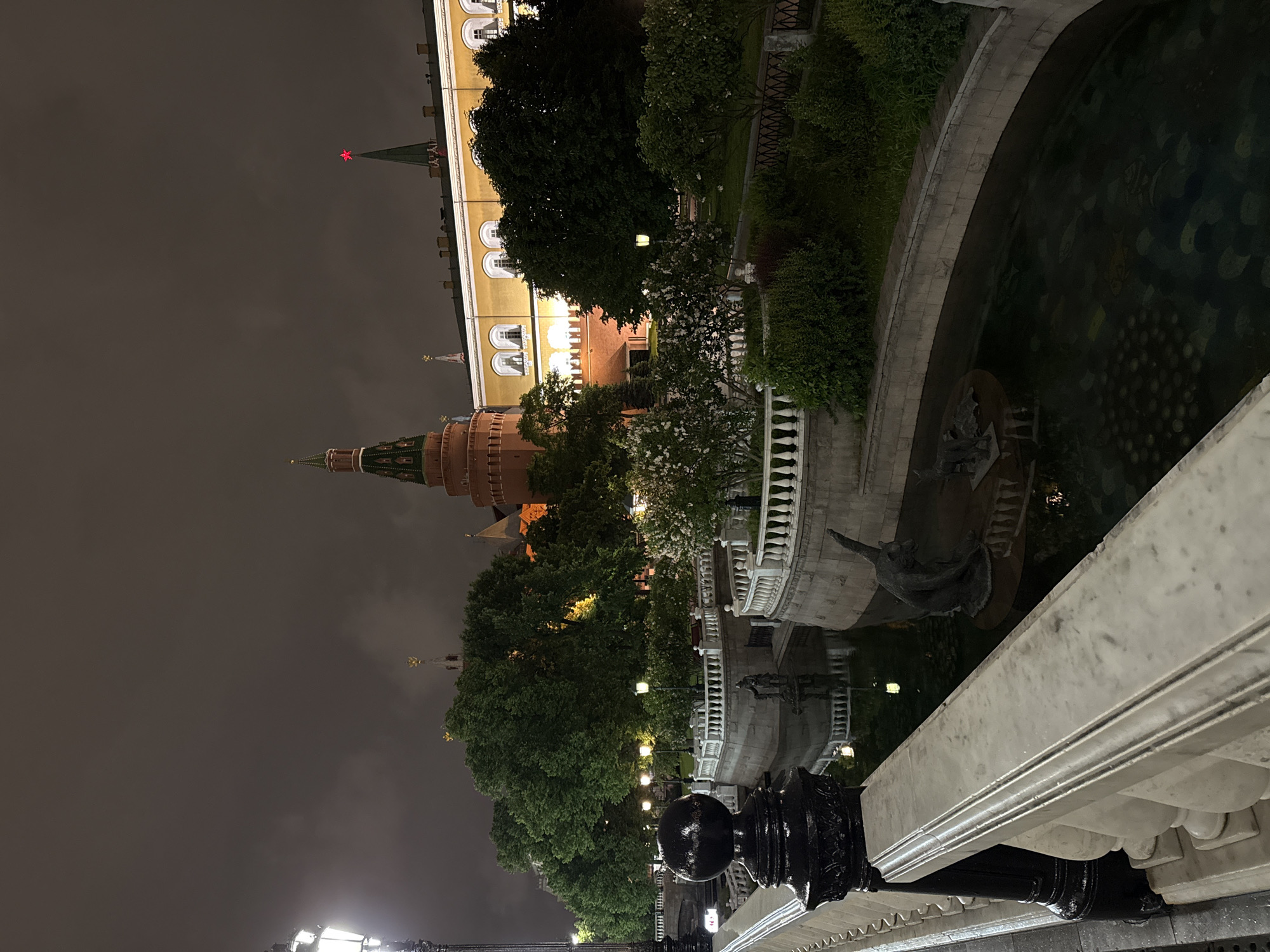 Captured from a low angle, the nighttime scene focuses on the Moscow Kremlin, distinguished by its iconic towers and golden-hued walls against a dark, overcast sky. The prominent tower in the foreground features a pointed, green roof. Part of the Kremlin's wall is illuminated, showcasing architectural details and windows.
Adjacent to the Kremlin, there's a park with mature trees and manicured greenery. A marble railing, complete with decorative black metal posts, defines the foreground perspective, suggesting a pedestrian pathway or bridge. A section of a water feature, potentially a pond or fountain, is visible with some sculpture in it.
The overall impression is one of a historic urban landscape viewed from a public space at night.