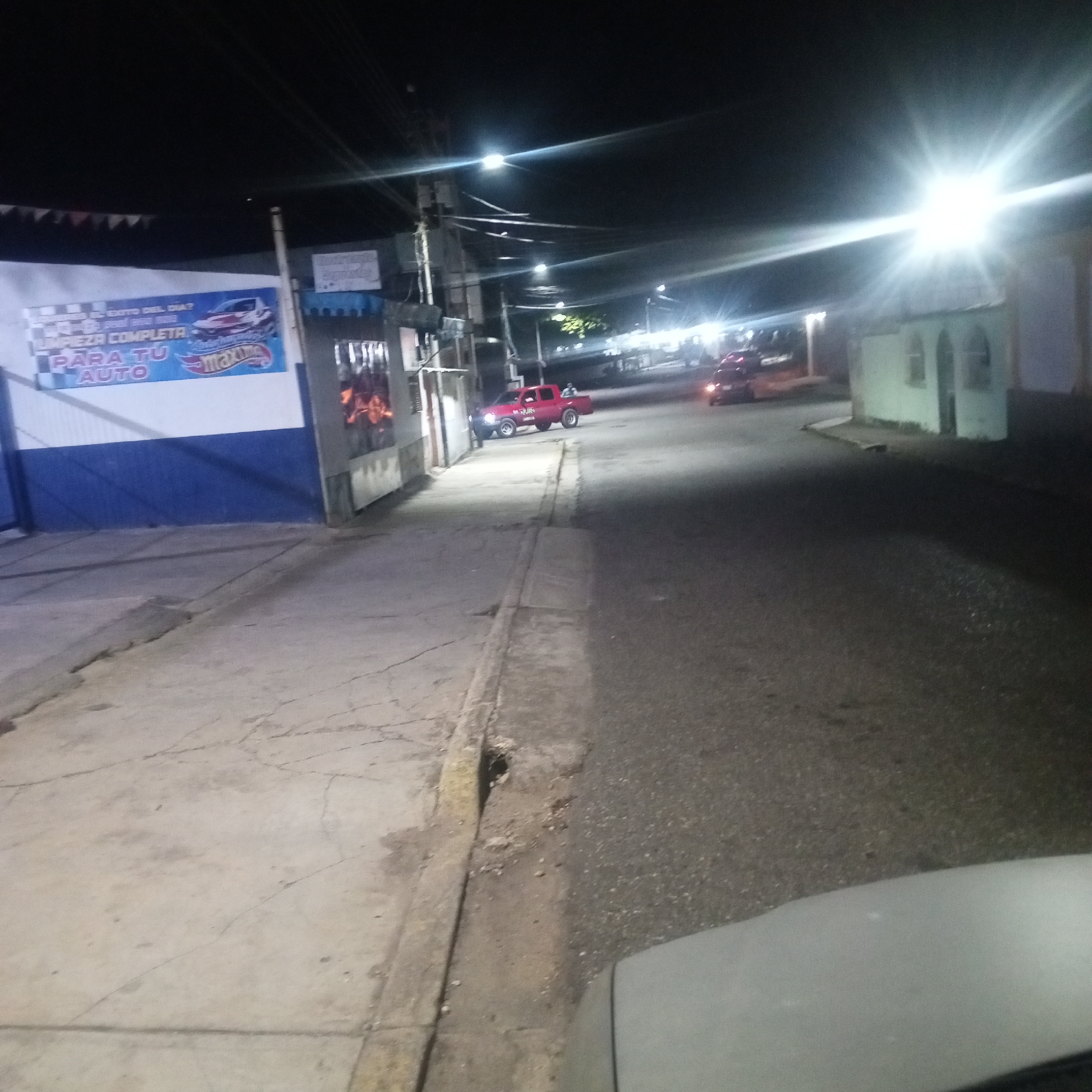 The image shows a street scene at night in Tinaquillo, Venezuela. The scene includes a building on the left with a blue and white sign advertising car wash services ("LIMPIEZA COMPLETA PARA TU AUTO"). A sidewalk runs along the building. Across the street, a red pickup truck is parked near the curb. Further down the street, another vehicle is visible, along with buildings on both sides of the road. Streetlights illuminate the scene, with one light on the right side appearing particularly bright. The bottom right corner of the image shows part of a car's hood.