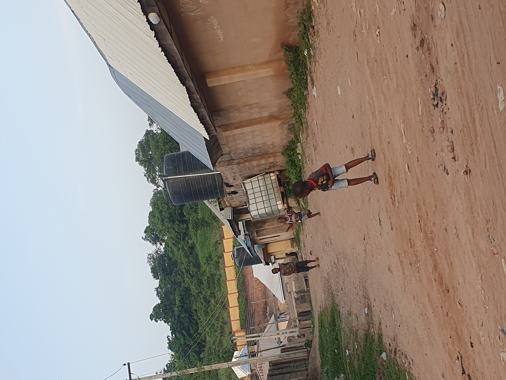 A wide shot captures a dusty street in Lokoja, Nigeria, with children playing. In the foreground, a young boy in a t-shirt and shorts stands with his back to the camera, facing a building. Further down the street, another child runs towards the right. In the mid-ground, a boy in a patterned t-shirt and shorts walks away from the camera. Behind the children, buildings with corrugated iron roofs line the street. Water tanks are visible on top of some buildings. A wooded hill forms the backdrop, with a yellow barrier fence running along its base. The sky is clear and overcast.