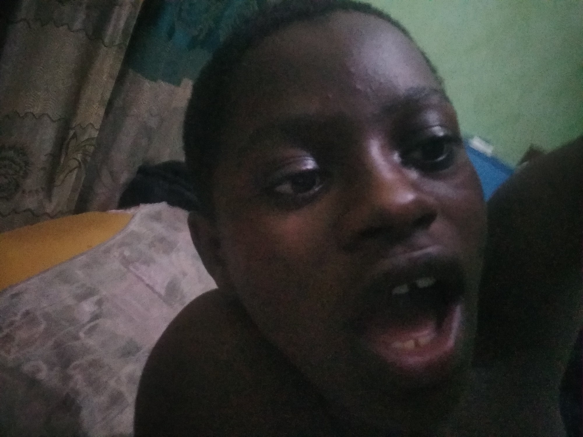 A dark-skinned person is the primary subject of the image, with their mouth open and teeth visible. The person is lying on a bed, with a patterned sheet visible. A wall is in the background. The location is the city of Shangisha, Nigeria.