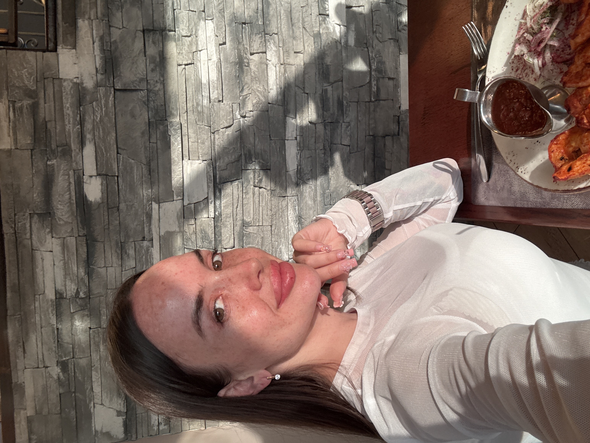 A woman is captured in a selfie at a restaurant. She has long, dark hair, brown eyes, and fair skin with freckles. Her lips are full and appear to be enhanced. She is wearing a white, sheer long-sleeved top with a high neckline and a silver watch on her left wrist. Her right hand is resting on her chin, and her fingernails are painted pink with nail art. She is wearing a small, sparkling earring in her left ear. In front of her, on a wooden table, is a plate of food that includes what appears to be grilled meat, a small metal dish of red sauce, and a side salad with onions. A fork and knife are also on the table. The background consists of a stone wall with a rough texture. Sunlight casts shadows on the wall, suggesting it is either early morning or late afternoon. The overall lighting is warm, with a strong source of light coming from the left. There is no visible indication of the specific city or country within the image itself, nor any landmarks that would definitively place it in Saint Petersburg, Russia.