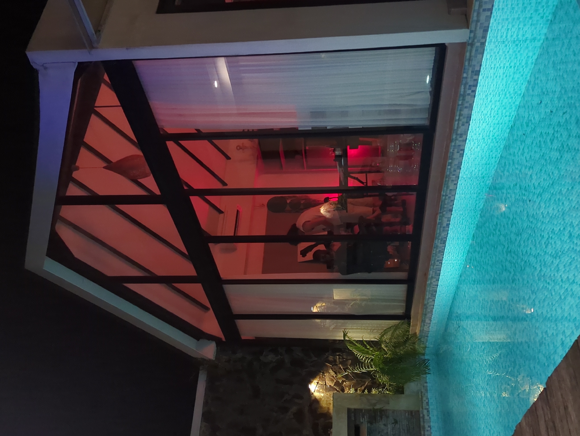 The image depicts the exterior of a building in what seems like Kediri, Indonesia, based on possible regional architectural styles. A section of the building has a dark-framed glass structure, possibly an enclosed patio or part of a restaurant, revealing an interior with red lighting, chairs, tables, and what appear to be people inside. The building has a white awning or overhang on the side of the glass enclosure. To the right of the enclosure, there's a swimming pool with blue-tiled edges and illuminated water. Below the glass enclosure is an area with a stone wall, plants, and what looks like a wooden structure. The surrounding area appears to be dark, suggesting it is night.