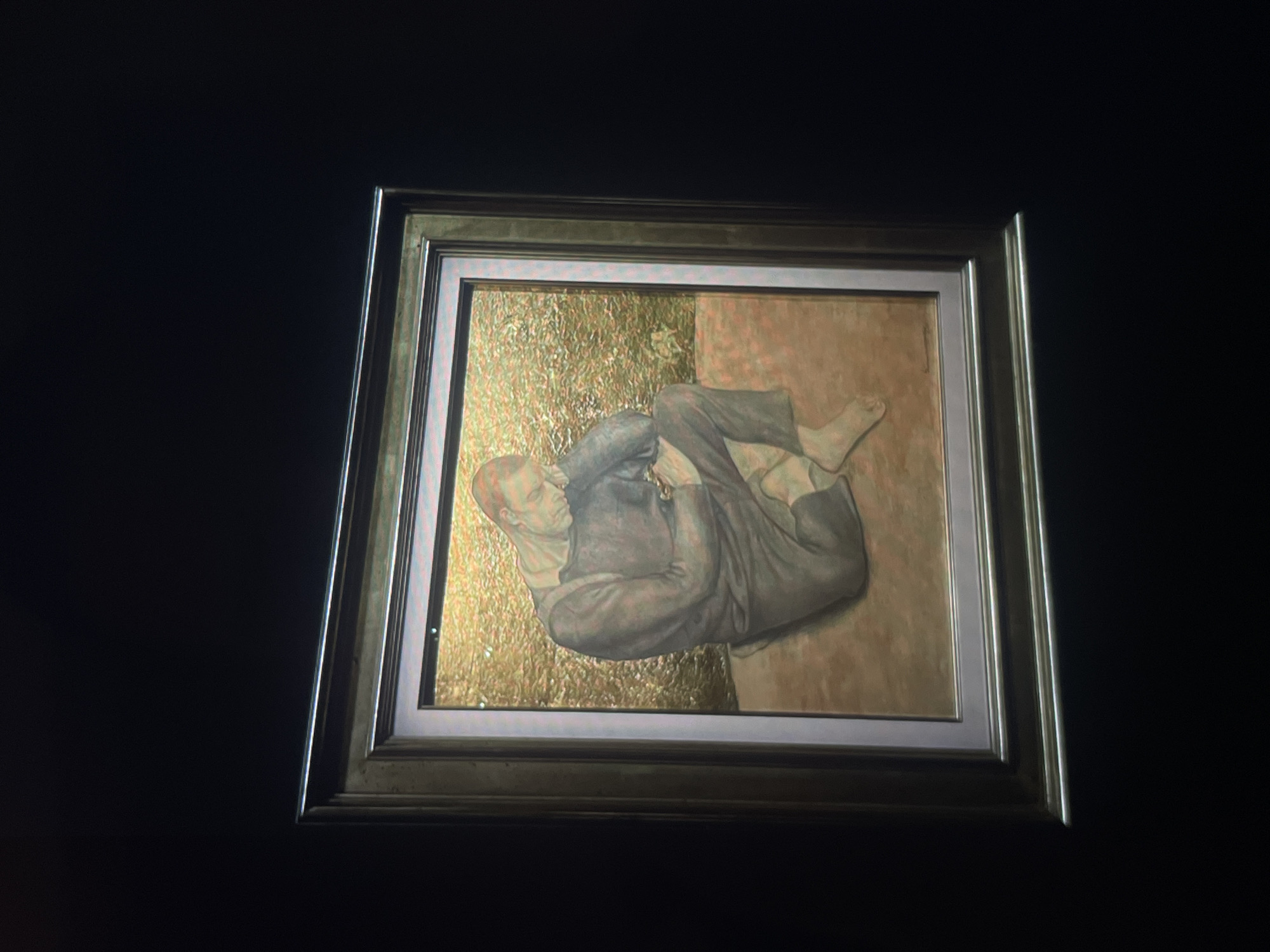 A framed painting is affixed to a wall of unknown color. The painting depicts a man reclining with legs crossed, dressed in a suit. The background of the painting is divided into two sections: the lower portion is textured gold, while the upper portion is a peach color. The painting is enclosed in a silver and bronze frame. The location is Unknown, Unknown.