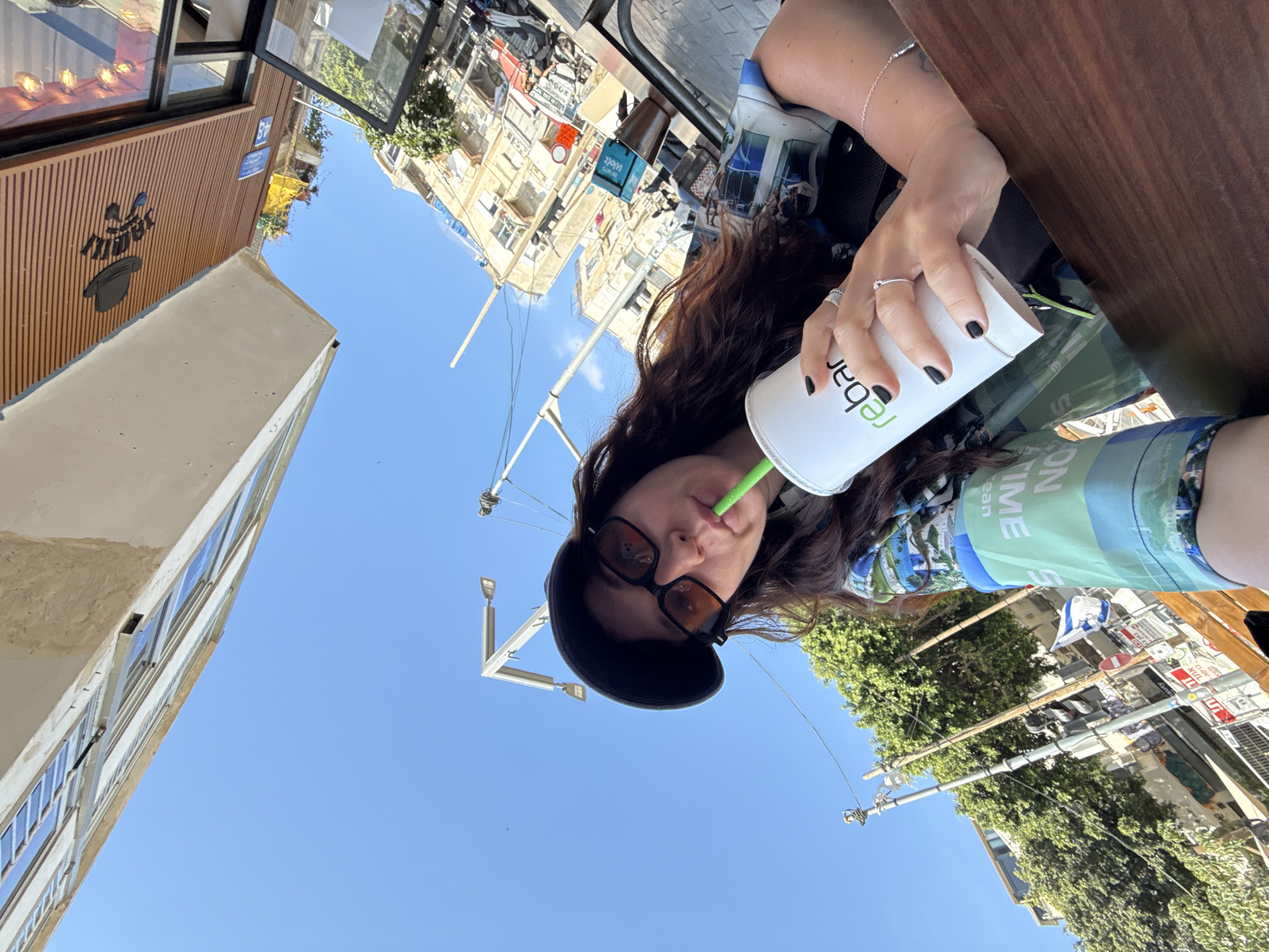 The image is taken from a low angle, capturing a woman holding a Rebar-branded drink with a green straw. She wears a black beret and sunglasses, and her fingernails are painted black. The background reveals an urban setting under a clear blue sky. Buildings with architectural details are visible, along with utility poles and trees lining the street. The location is likely Tel Aviv, Israel, based on the street signs written in Hebrew and possible Israeli flags shown.