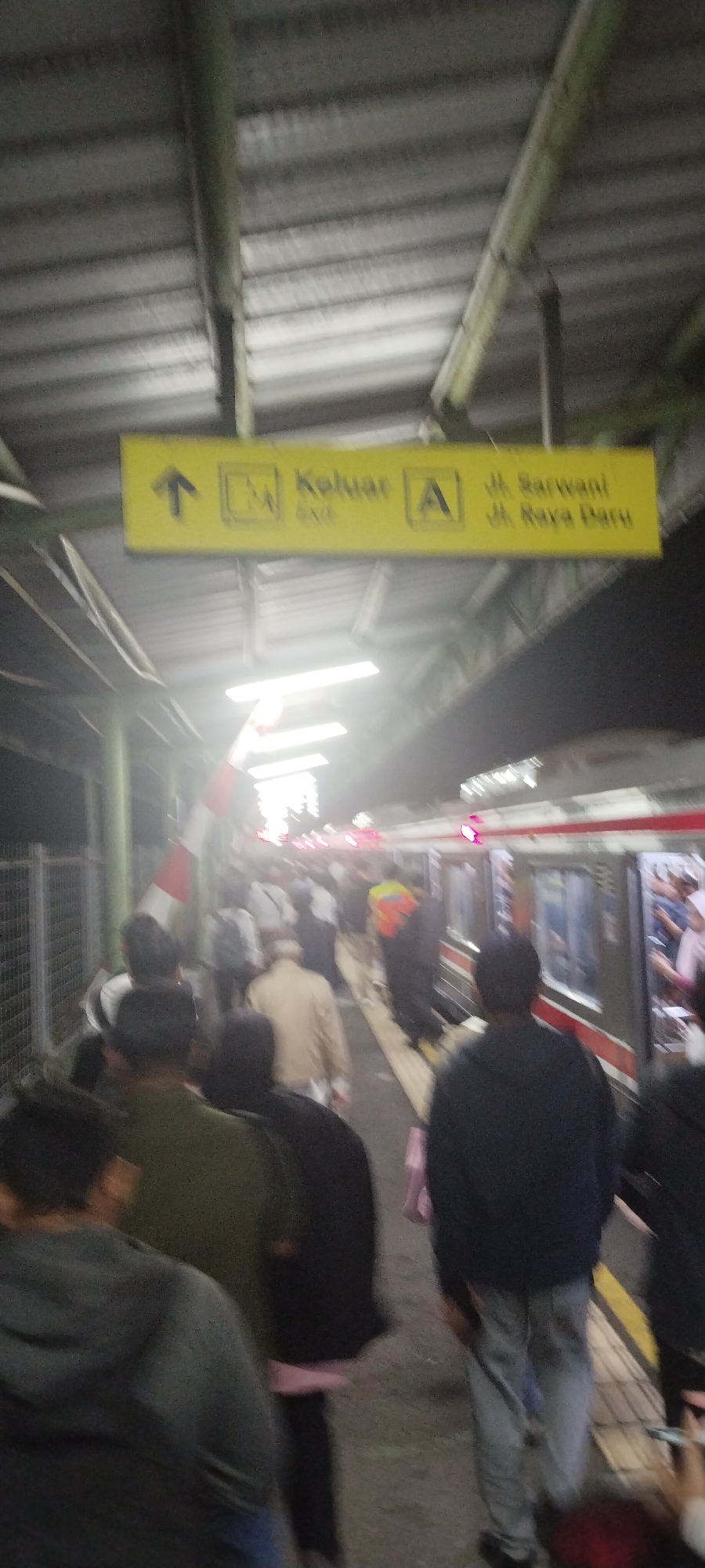 The image shows a blurry view of a train station platform in Daru, Indonesia. A yellow sign indicates "Keluar A" (Exit A) and street names "Jl. Sarwani" and "Jl. Raya Baru". A train is visible on the right side of the image. Many people are on the platform, appearing to be waiting for or disembarking from the train. The platform is covered by a corrugated metal roof supported by pillars. Lighting is provided by overhead fluorescent lights. The scene depicts a busy public transportation hub.