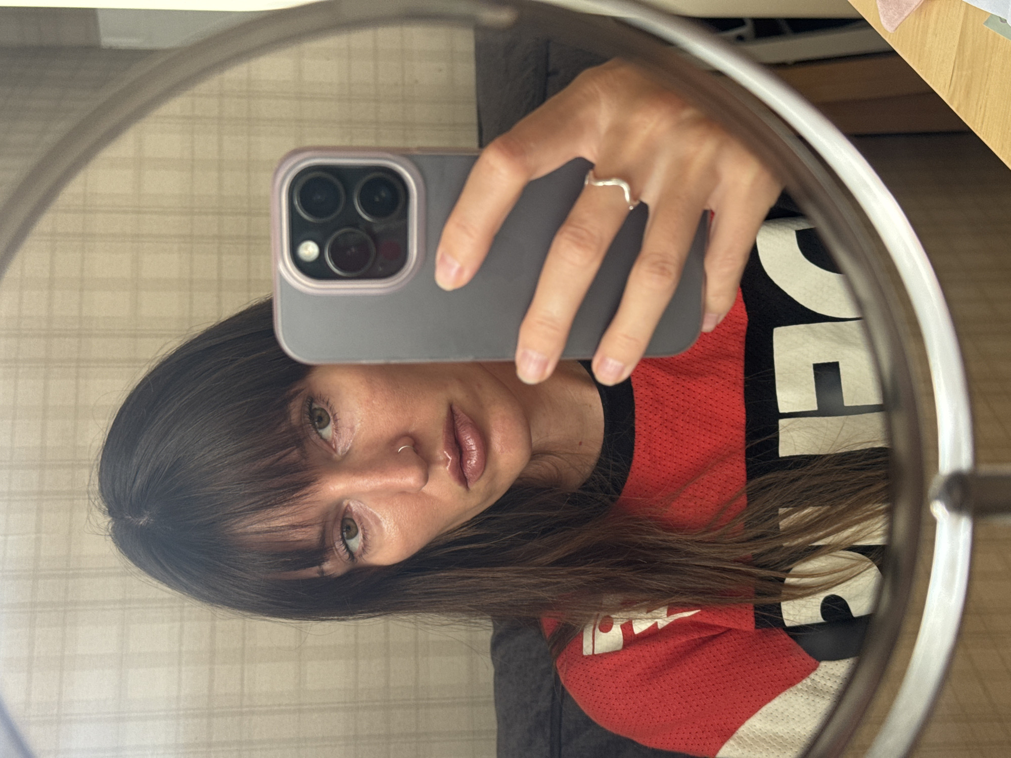 A woman with long, dark hair and bangs is taking a mirror selfie. She is wearing a red and white long-sleeved shirt with black lettering. She is holding a grey phone with a purple case in her right hand. Her left hand, wearing a silver ring, is visible on the phone. The background appears to be a wall with a plaid pattern. The image is cropped and framed by a circular mirror. The location is not specified in the image or the prompt.