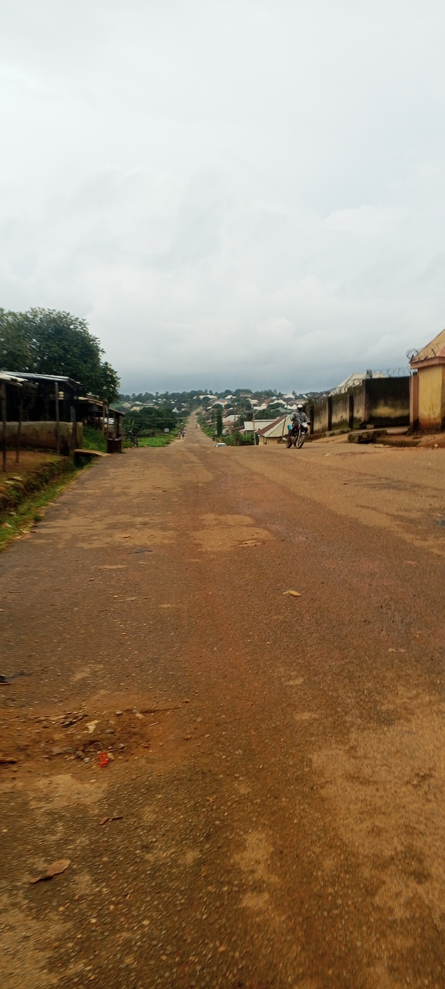 The image depicts a rural road in Bahit, Nigeria, under an overcast sky. The road is unpaved and appears to be made of dirt and gravel, with some patchy areas of asphalt. The road stretches into the distance, leading uphill towards a collection of buildings that make up a village or small town. On either side of the road are various structures, including some with corrugated metal roofs and others that appear to be more rudimentary. There is a dense cluster of green trees on the left side of the road, partially obscuring some of the buildings. In the distance, a scattering of houses and buildings are visible on the rolling hills. A motorcycle with two people on it is traveling up the road on the right side. A single white car is also visible further up the road. The foreground of the image shows the texture of the road surface with debris scattered across it.