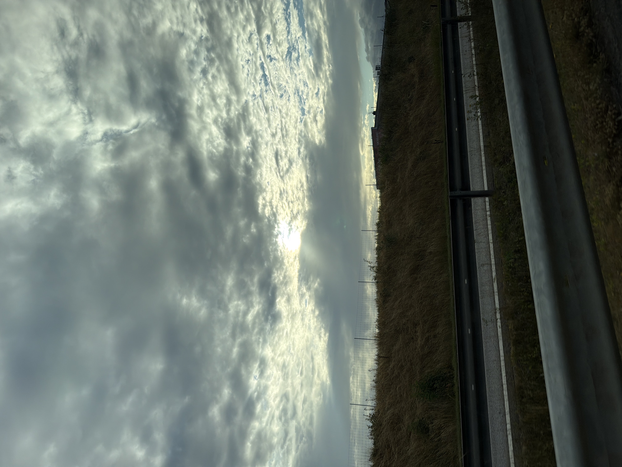 The image shows a cloudy sky with sunlight breaking through in the upper central part of the frame. Below the sky, there is a grassy embankment with a chain-link fence running horizontally across it. Beyond the fence, a building or structure is visible on the horizon. In the foreground, there is a road with guardrails. The scene appears to be along a highway in Tofta, Sweden, based on the general landscape and the presence of typical Swedish roadside infrastructure. There are no people or animals visible in the image.