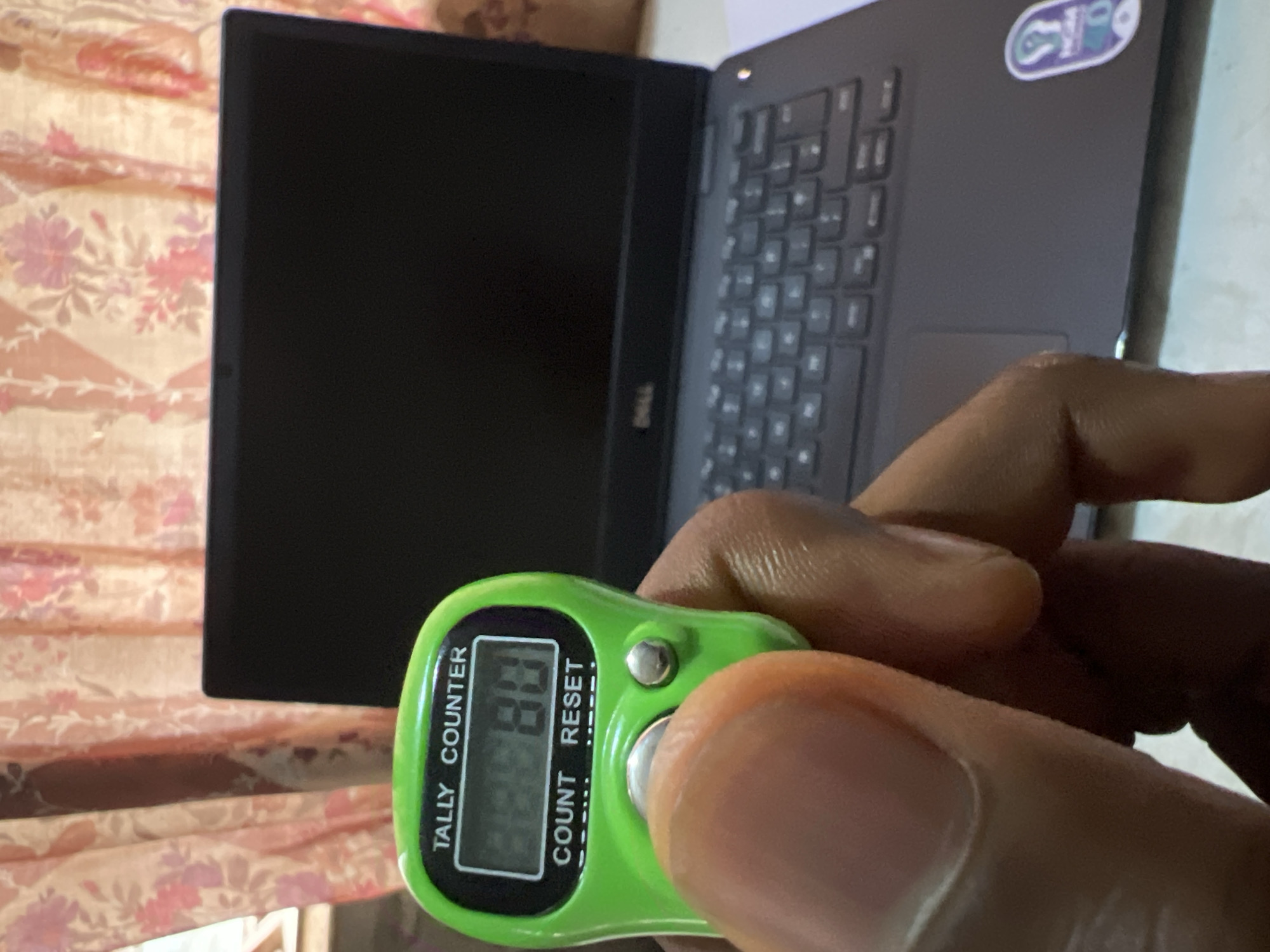 A person's hand holds a green digital tally counter displaying the number "80," with a black Dell laptop in the background. The background includes a floral-patterned curtain or fabric. The scene is likely indoors, possibly in a home or office environment. No specific location within Minna, Nigeria, can be determined.