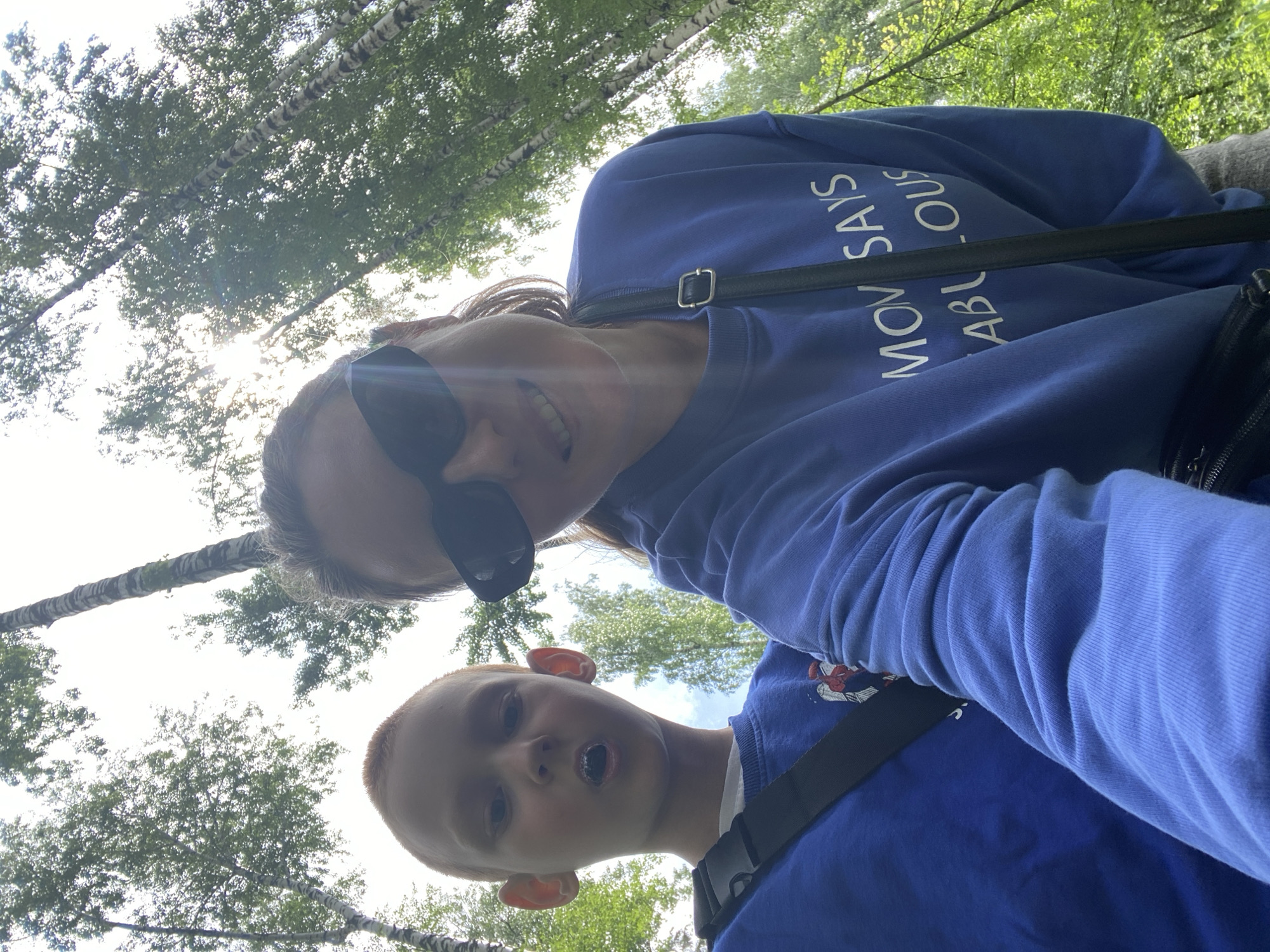 Two people, a young boy and an adult female, are captured in a low-angle, selfie-style photograph taken outdoors amidst a forest of birch trees. The sun is visible through the canopy, creating lens flare.
The boy, positioned on the left, has fair skin, reddish hair, and appears to be making a surprised expression with his mouth open, revealing white teeth or an object inside his mouth. He is wearing a blue sweatshirt with a graphic of a character, possibly from a comic book, on the left side of his chest. A black strap is diagonally across his chest.
The adult female is on the right, wearing large dark sunglasses and a blue sweatshirt with white lettering that partially reads "MOM SAYS FABULOUS." She has dark hair pulled back. A black purse or bag with a strap is visible, crossing her chest and resting on her shoulder. The background consists of tall, slender birch trees with white bark and green foliage, suggesting a deciduous forest. The sky is bright, indicating daytime. The context of Noviy, Russia, is not directly evident from visual cues within the image itself but is provided as location information.