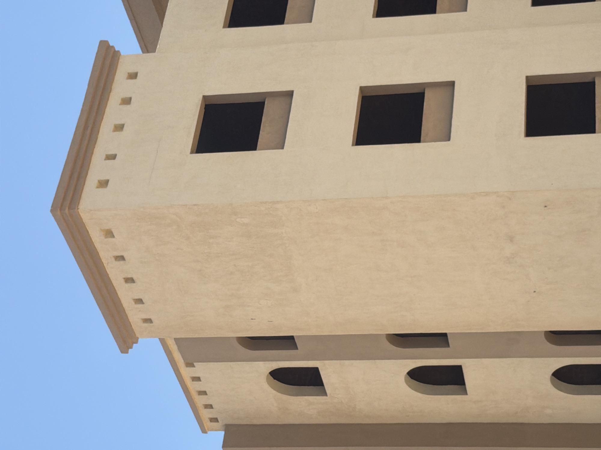 This is a close-up, eye-level, outdoor, daylight, vertical, medium shot of a tan, stucco building under a clear blue sky. The building appears to be under construction or renovation, as it has many unglazed window openings. The building has a modern architectural style with clean lines and a flat roof. The facade features a repeating pattern of rectangular openings on the upper floors and arched openings on the lower floors. The building is situated in a city, but no specific location can be determined from the image. The image is devoid of any people or specific cultural markers.