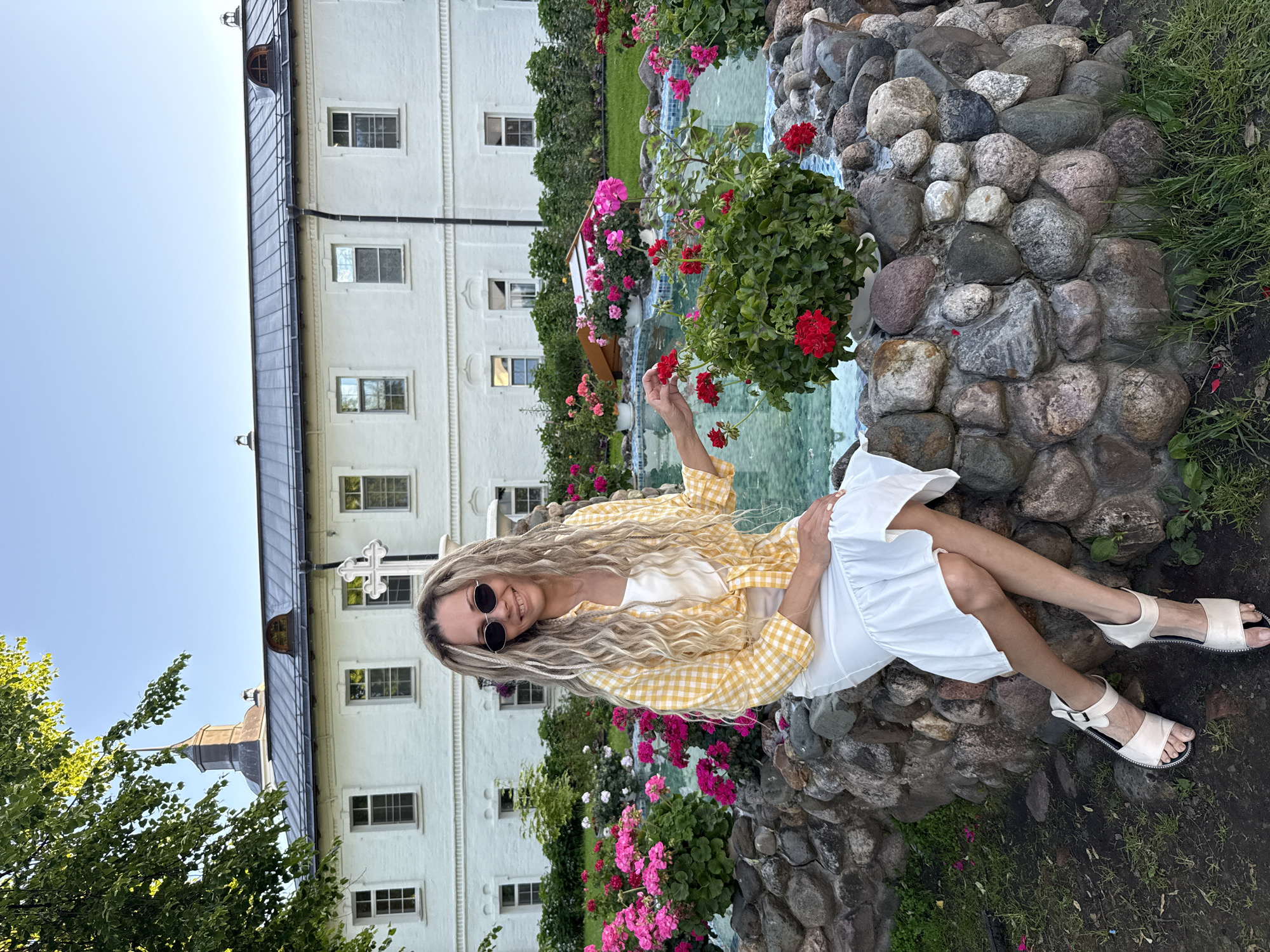 The image depicts a woman seated on the stone edge of a water feature in a garden, with a large, white architectural building in the background. The location is identified as Yaroslavl, Russia.
The woman has long, blonde, dreadlock-style hair and is wearing sunglasses with oval frames. She is smiling, looking towards the viewer. Her attire consists of a white tank top, a yellow and white gingham-patterned long-sleeved shirt tied at the waist, and a long, white, tiered skirt. She wears light-colored flat sandals. Her right hand is gently touching a red flower in a pot.
The water feature, a pond or fountain, has edges constructed from diverse rounded natural stones. Its interior basin appears to be tiled in light blue, visible through the clear water. Several potted plants, predominantly red geraniums, are placed on the stone edge of the fountain. Behind the fountain, extensive flowerbeds feature vibrant pink blossoms, with additional greenery and bushes forming a hedge.
The building in the background is a multi-story, white structure with a dark, possibly metal, roof. It features numerous rectangular windows with white frames. A prominent white decorative cross on a pedestal stands in front of the building's facade, slightly left of center. Above the left portion of the building, a dark cupola with a pointed spire is visible. The sky is clear and light blue, suggesting fair weather and late afternoon or early evening lighting. Green grass is visible in the foreground and a tree branch with green leaves is partially seen in the upper left corner.