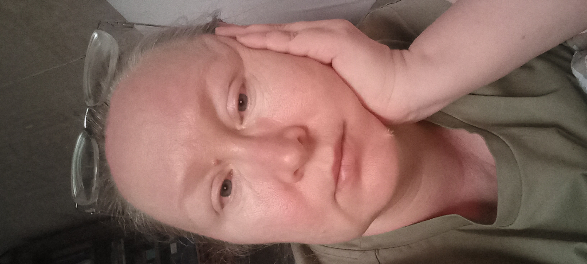 A woman is seen in a close-up, head-and-shoulders shot, resting her right cheek on her right hand. She is wearing a green shirt with a wide neckline. Her eyeglasses are pushed up onto her forehead, revealing her forehead, eyes, nose, and mouth. The background is dark and appears to be indoors, possibly a home environment, with what looks like bookshelves on the left. There is no indication of the city of Izhevsk, Russia, in the image.