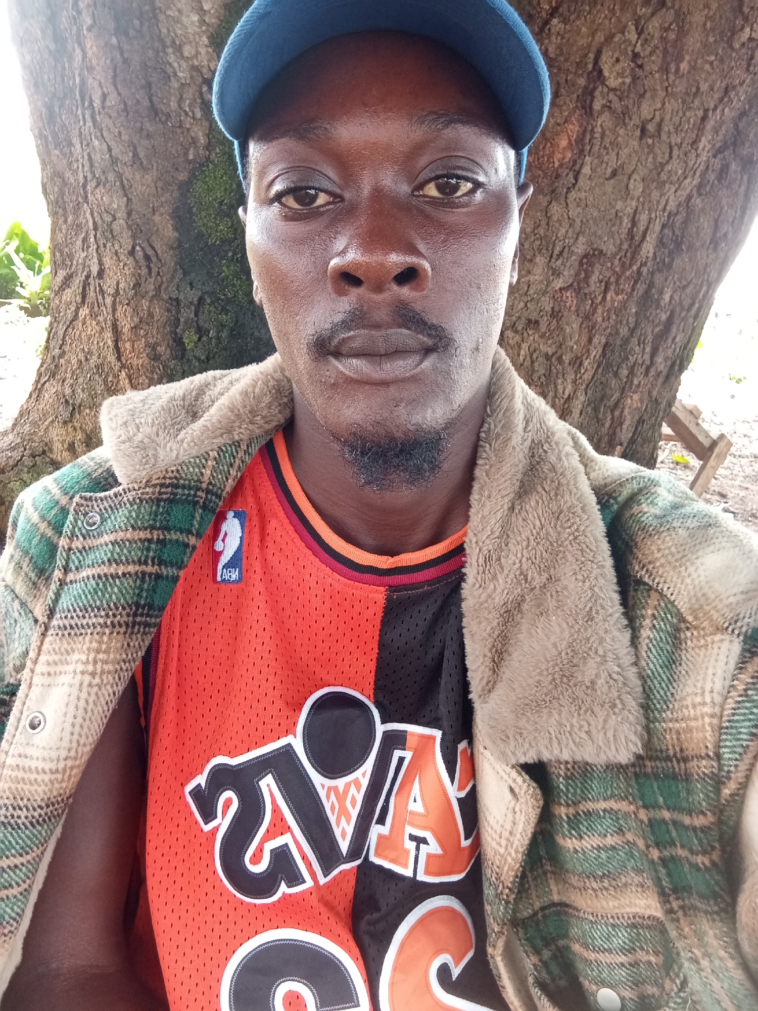 In Fadan Kuower, Nigeria, a young man with dark skin, a mustache, and a goatee, wearing a blue cap, a fuzzy collared plaid jacket over an orange and black basketball jersey, poses for a selfie. He stands in front of a large tree. The background shows a wooden structure and foliage.