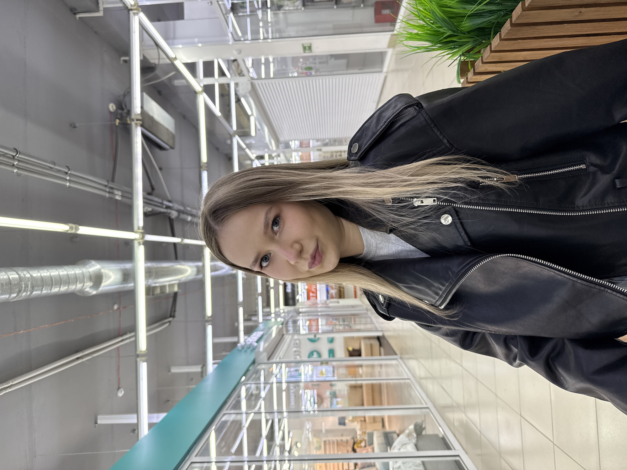 A young woman stands indoors, possibly in a shopping center or large store, judging by the ceiling lighting and storefronts visible in the background. She is wearing a black leather jacket over a gray top. The environment features industrial-style overhead piping and framework, with storefronts and merchandise displays in the background. The location is the city of Chaikovsky, Russia.