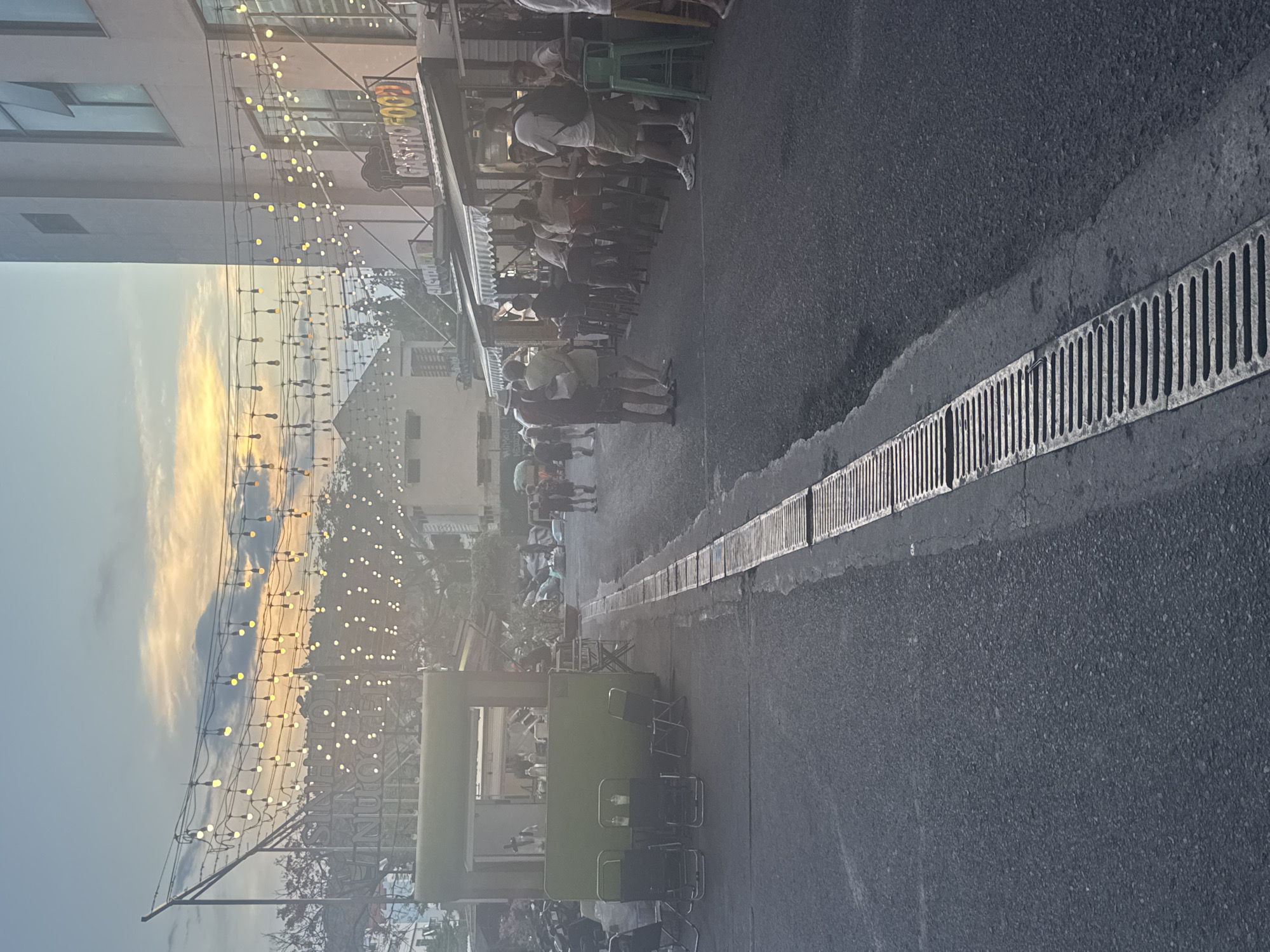 The image depicts a street scene at dusk, likely in Bac Nha Trang Ward, Vietnam, characterized by a warm sky with clouds and illuminated by strings of bare light bulbs overhead. The street is paved with asphalt, featuring a prominent drainage grate running along the right side.
On the left, a small, green food stall or kiosk is visible, with a counter and what appear to be two chairs and tables set up outside, suggesting a casual dining area. Beyond the stall, motorcycles are parked.
Further down the street, to the right, several businesses with awnings line the road. One sign reads "GASTRO FOOD." People are gathered at tables in front of these establishments, some seated and some standing, engaged in social interaction. The atmosphere appears to be relaxed and communal, with the overhead lights creating a festive ambiance. The overall scene suggests a lively evening street market or dining area.