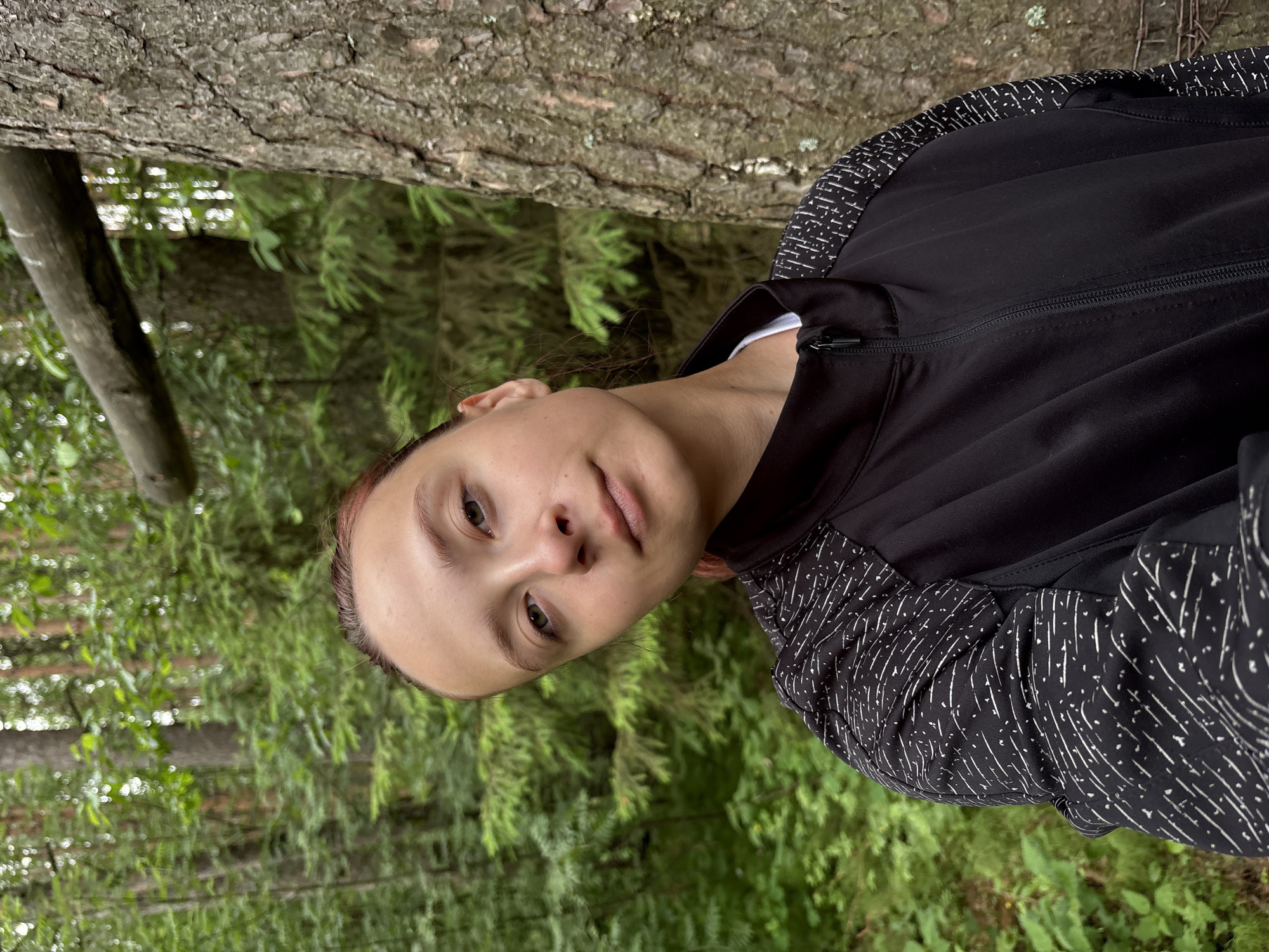The image, rotated 90 degrees counter-clockwise, depicts a young person positioned outdoors in a forest setting. The individual has light skin, dark hair with subtle reddish undertones, and light-colored eyes, and is looking directly at the camera with a neutral and calm expression.
They are wearing a black athletic jacket with a visible zipper down the front. The jacket features a distinctive pattern of short white dashes and dots on the shoulders and upper sleeves, providing a contrast to the solid black body. A white collar or undershirt is visible beneath the jacket.
The background is a dense, out-of-focus forest, filled with lush green foliage and numerous tree trunks, suggesting a natural and secluded environment. A large tree trunk with textured bark is prominent to the upper left of the person. The lighting appears natural and diffused, indicating daytime. The vibrant green leaves suggest a warm season. This type of wooded landscape is consistent with natural areas found in regions like Chaikovsky, Russia. No visible text is present in the image.