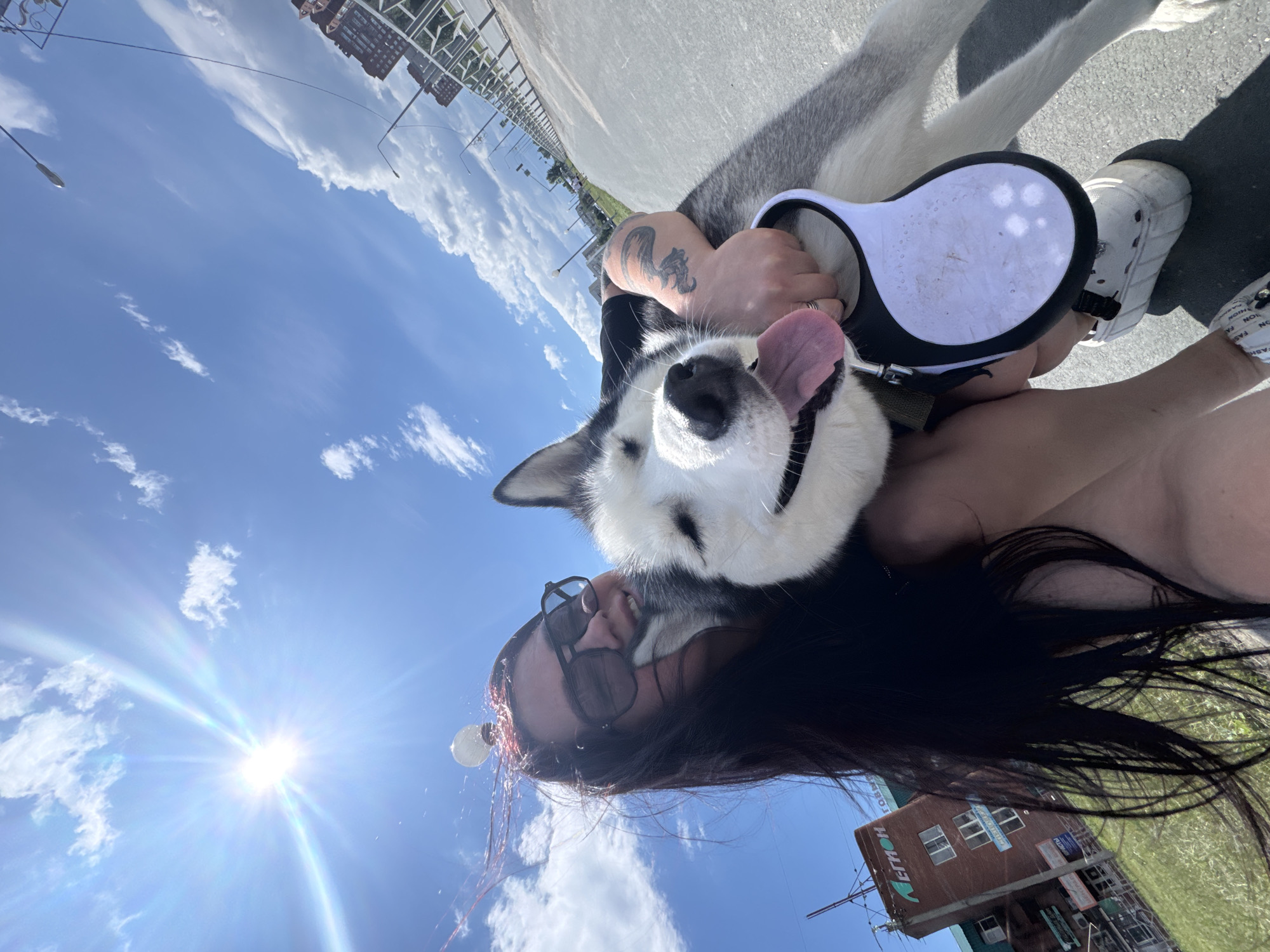 In an outdoor, urban setting under a bright, sunny sky with scattered clouds, a person with long dark hair and sunglasses is embracing a husky dog. The person is smiling, and the dog has its tongue out, appearing happy. The person is wearing a black shirt and a bracelet. The husky is wearing a collar and is attached to a retractable leash, which the person is holding. The background shows a paved walkway, a grassy area, a brick building with signage, and residential buildings in the distance. Streetlights are visible along the walkway. The image is taken from a low angle, looking up towards the sky.