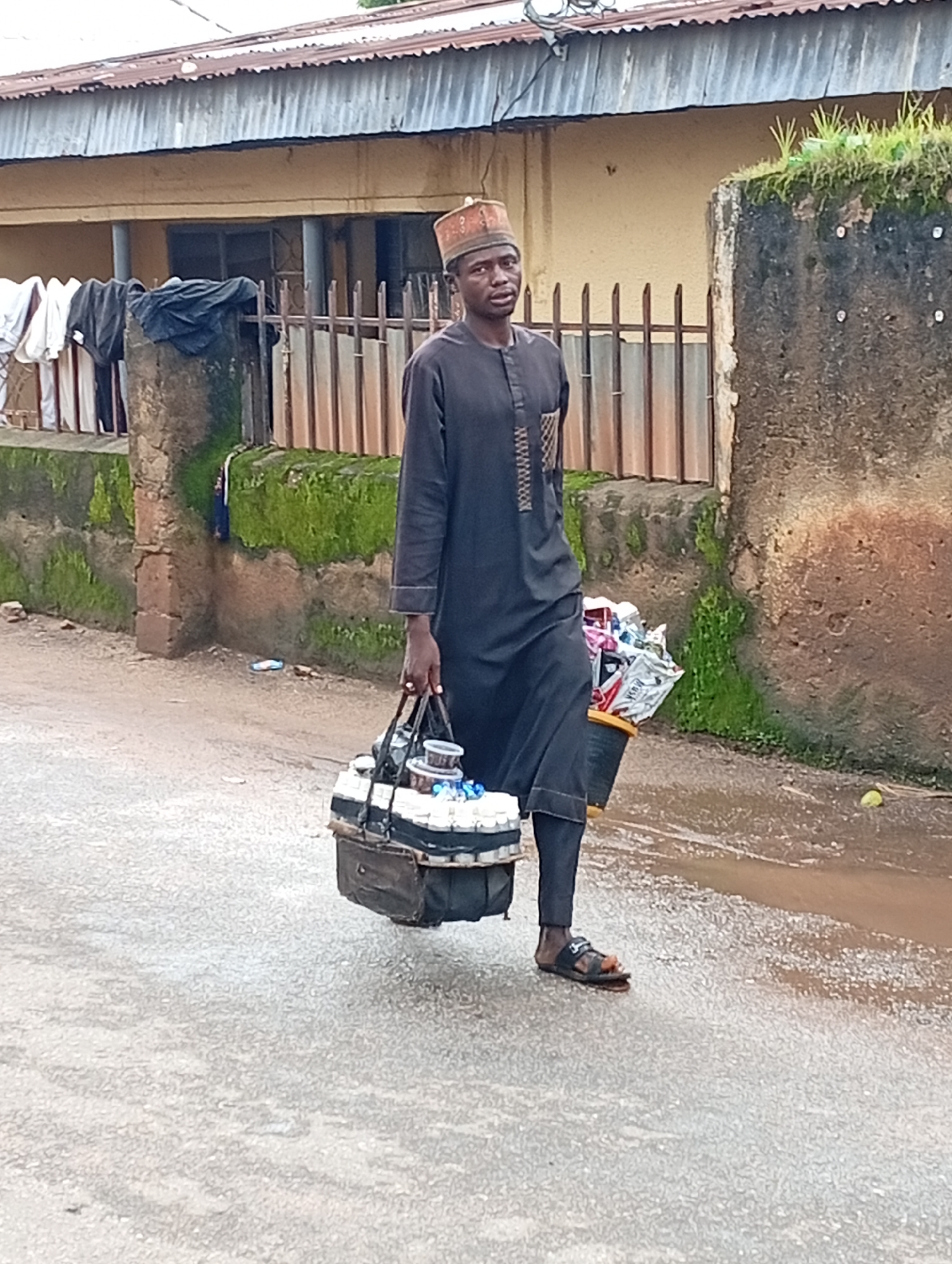 In a street scene in Jos, Nigeria, a young man walks on a wet asphalt road. He is dressed in a dark, long-sleeved tunic with white trim and wears a patterned cap. In his right hand, he carries a large black bag that appears to be a cooler, filled with multiple white bottles. To his right, he is carrying a yellow bucket containing various items wrapped in newspaper. The background shows a tan building with a corrugated iron roof, a metal fence, and a moss-covered wall. To the left, laundry is hanging on a line attached to a stone pillar. The ground is wet, indicating recent rain.