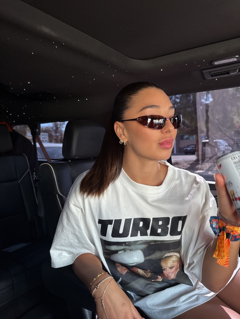 A young woman is depicted from the chest up, seated inside a car during clear daytime conditions. She has dark hair styled in a sleek, pulled-back ponytail and wears dark, narrow-framed sunglasses. Her lips are painted with a light pink or nude lipstick, and she has several small gold hoop and stud earrings in her left ear.
She is wearing a loose-fitting white t-shirt featuring a prominent graphic print. The print includes the bold, black word "TURBO" at the top, beneath which is a vintage-style image of two individuals (likely women) looking upwards, as if reflected in a car's rear-view mirror or seen through a sunroof. On her left hand, she sports a delicate, layered gold bracelet that extends to a ring on one finger. On her right wrist, she wears multiple colorful fabric wristbands, possibly from an event or festival. She holds a white can with colorful graphics and partial text, appearing to read "CELCIUS," in her right hand.
The car's interior features dark leather seats and a unique dark headliner embedded with numerous small white lights, creating a "starry night" effect. Bright sunlight illuminates the scene, casting shadows and indicating a sunny day. The background visible through the car windows is blurred but suggests an outdoor setting with trees and possibly other vehicles. The woman gazes slightly to her right (viewer's left), with a relaxed expression.