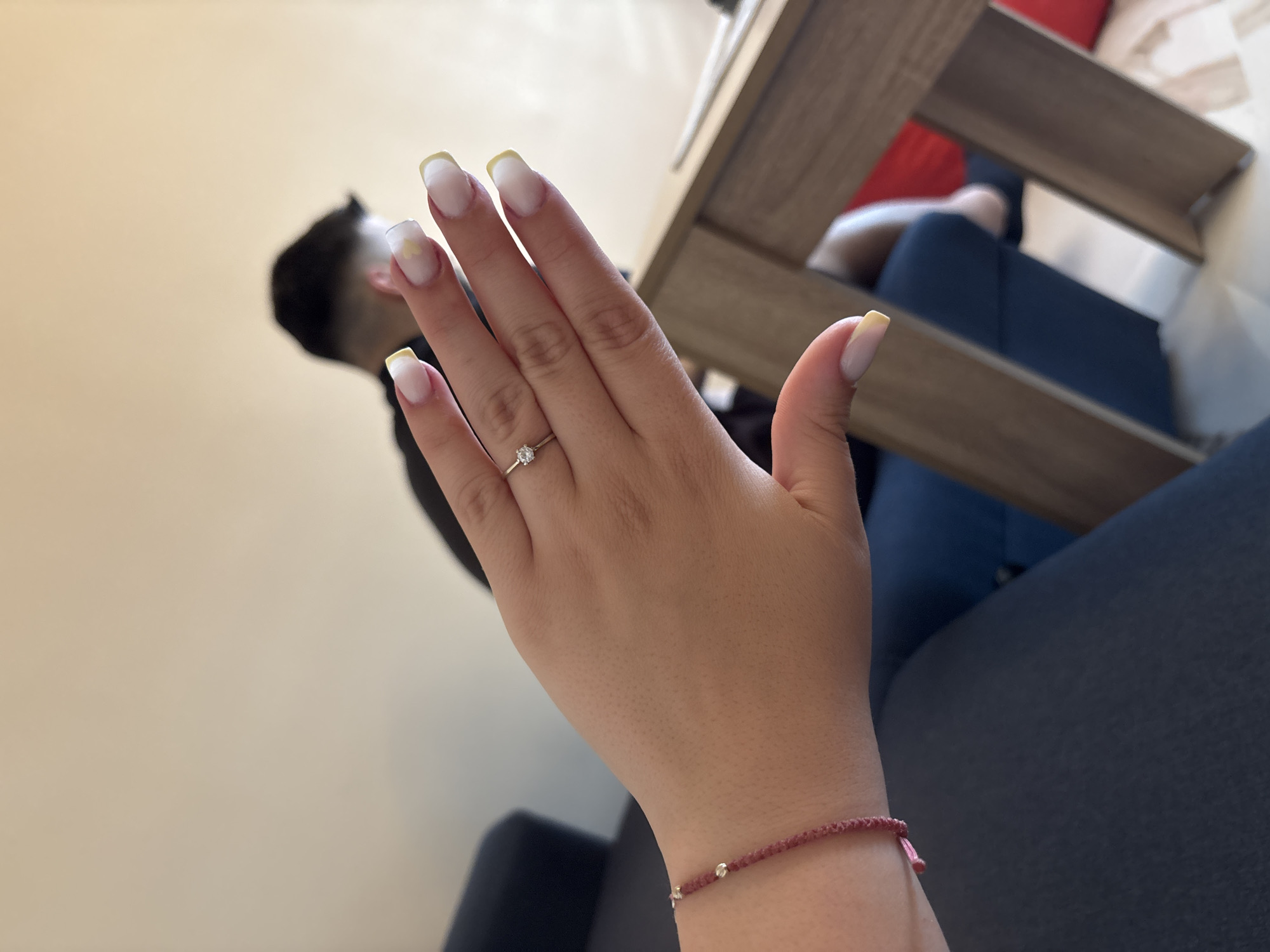 The image provides a close-up view of a person's left hand in the foreground, angled slightly upwards, against a blurred indoor background.
The hand has light-toned skin. On the ring finger, a silver-colored ring with a small, clear gemstone is worn. A thin, reddish-pink beaded bracelet with small silver-colored accents adorns the wrist. The fingernails are long and manicured, featuring a French tip style with white bases and bright yellow tips. A small yellow heart design is visible on the index finger's nail.
In the soft-focused background, the back of another person's head and shoulder are visible on the left, suggesting dark hair and dark clothing. To the right, a light brown wooden furniture piece, possibly a table or shelf, spans horizontally. Through and around this furniture, a dark blue fabric (likely clothing or upholstery) and a vibrant red fabric object are discernible. The wall behind is a plain, light color.
The scene appears to be an indoor setting, possibly a home or cafe, during daylight hours. The primary focus of the image is on the hand, specifically showcasing the ring and the manicure. There is no specific activity or event clearly depicted beyond this display. No text is visible in the image. While the location context of Varna, Bulgaria, is provided, no visual cues within the image itself confirm this.