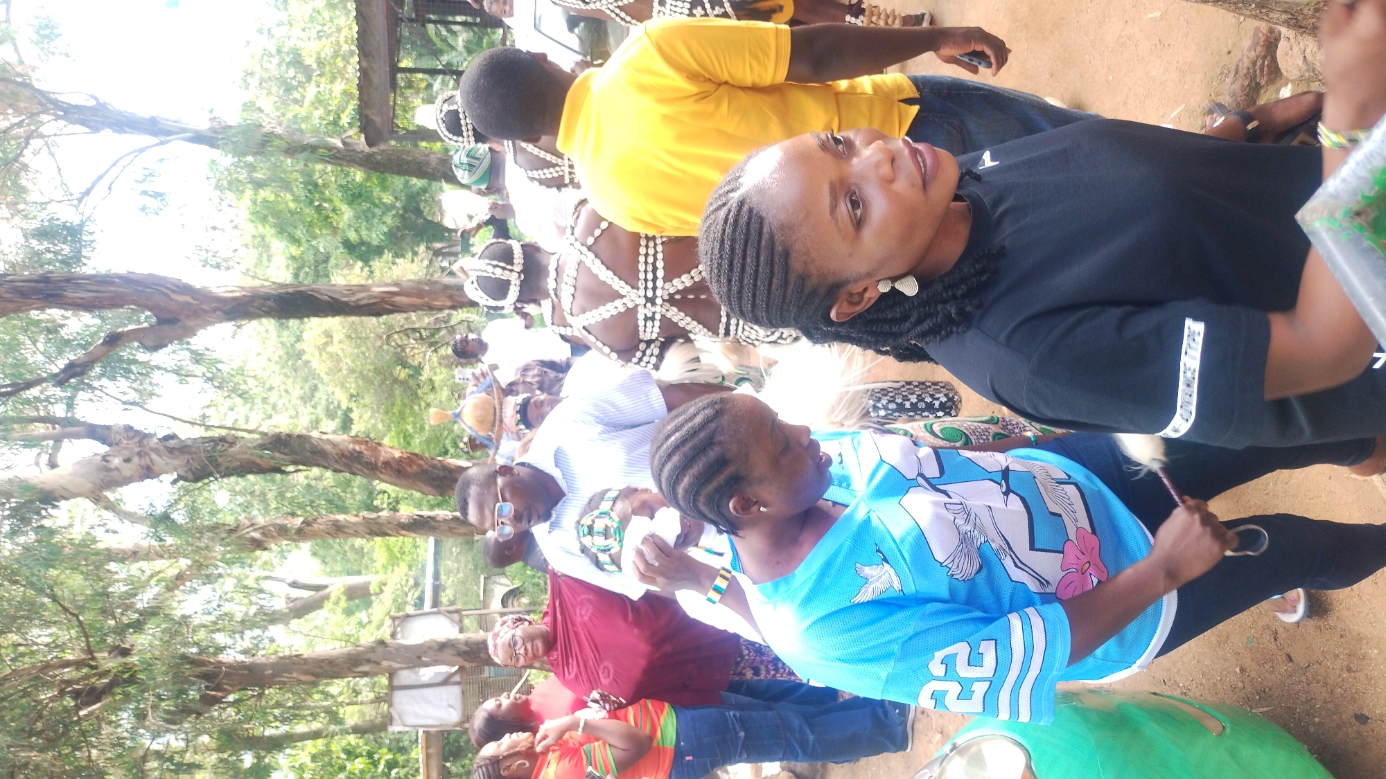 In Jos, Nigeria, a group of people are gathered outdoors, possibly for a cultural event or celebration. The scene is filled with lush greenery from trees, suggesting a park or a less urbanized area within the city. In the foreground, two women are prominent. One, on the left, wears a bright blue t-shirt with white and light blue graphics, including the number '22' on the sleeve. She is holding a small, fluffy whisk. The other woman, in the center-right, has dark braided hair and is wearing a black t-shirt with "ADVANCE TYPE" written on the sleeve. Her expression is attentive as she looks to the right.
Behind them, a man in a light blue striped shirt and glasses is visible, along with other individuals dressed in traditional attire adorned with shells, indicating a cultural performance or ceremony. One person in the background wears a vibrant yellow shirt. The overall atmosphere appears to be one of community and engagement with cultural traditions. The presence of a green vehicle on the left side of the frame suggests a public space accessible by road. The ground is a mix of dirt and sand.