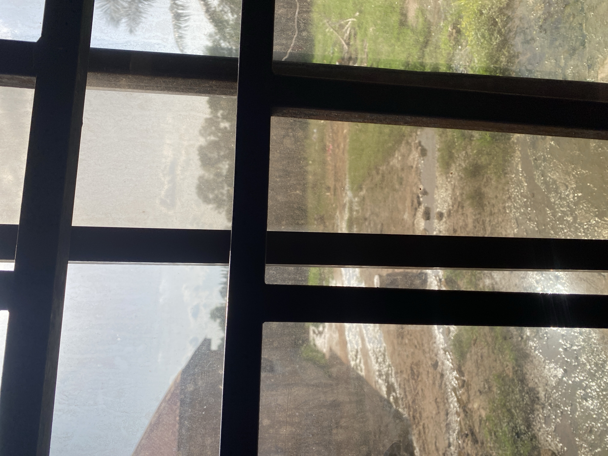 This image is a view through a window with dark metal bars, likely in Jauro Yinu, Nigeria. The windowpanes are somewhat obscured by dust or grime, creating a hazy effect. The scene outside suggests an outdoor environment, possibly rural.
In the upper left quadrant of the image, a portion of a palm frond is visible, hinting at the presence of tropical vegetation. The central and right portions of the image show a body of water, possibly a stream or shallow river, with sunlight glinting off its surface. The banks of the water appear muddy and uneven, with patches of green vegetation, indicating a natural landscape. Some debris or ripples are noticeable in the water.
The lighting suggests it is daytime, with bright sunlight illuminating the outdoor scene. The atmosphere appears to be somewhat hazy, which could be due to the dust on the window or the general weather conditions. There are no people or discernible human activities explicitly visible in the frame. The overall impression is one of a quiet, perhaps somewhat rustic, outdoor setting observed from indoors.