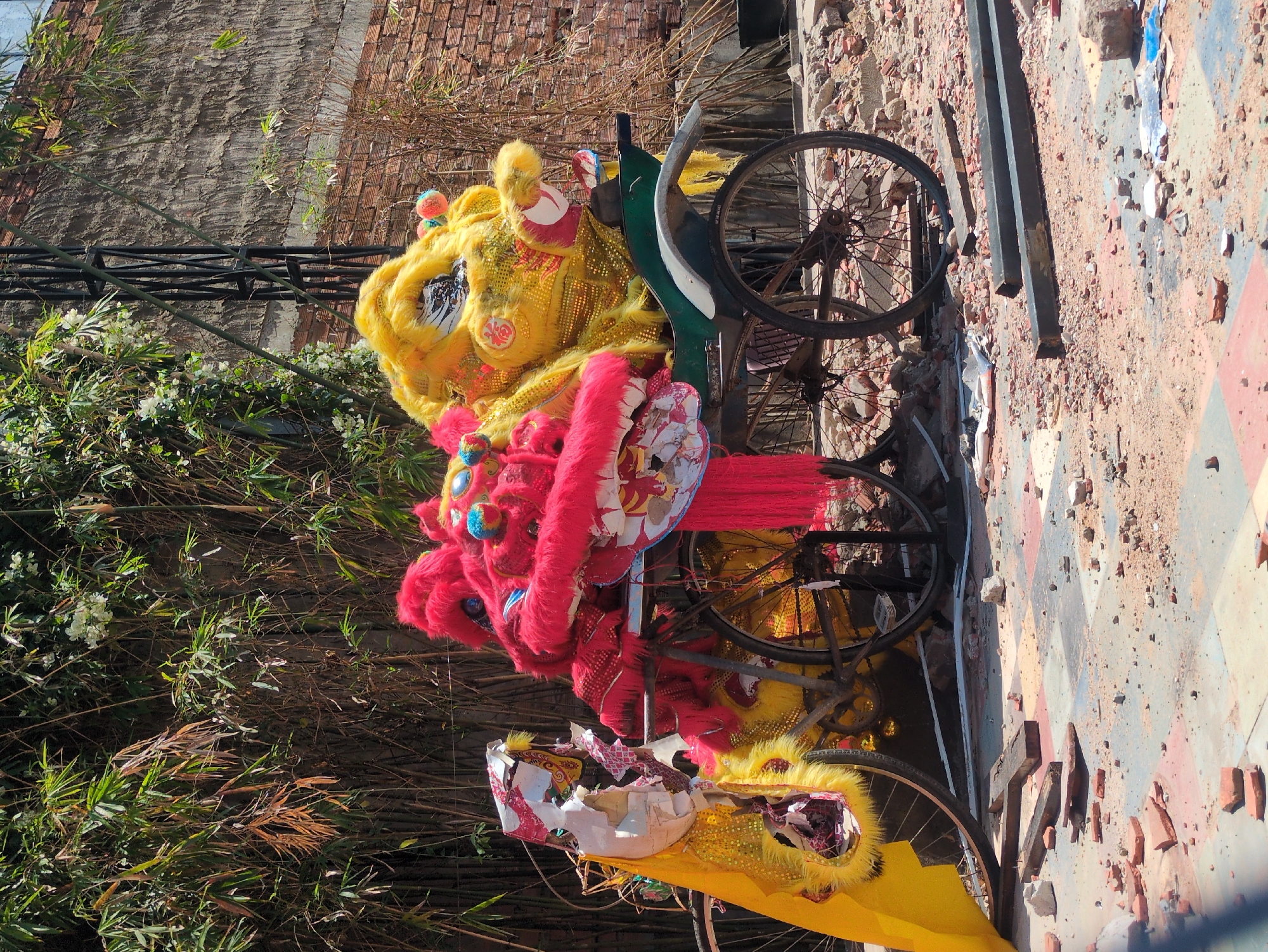 A rickshaw-like vehicle holds three lion dance costumes, in the city of Phú Quốc, Vietnam. The costumes are stacked one on top of the other, and are colored yellow, red, and white. The wheels of the vehicle sit upon a broken brick surface, and are surrounded by debris. Vegetation grows behind the vehicle, and a brick wall stands further in the background.