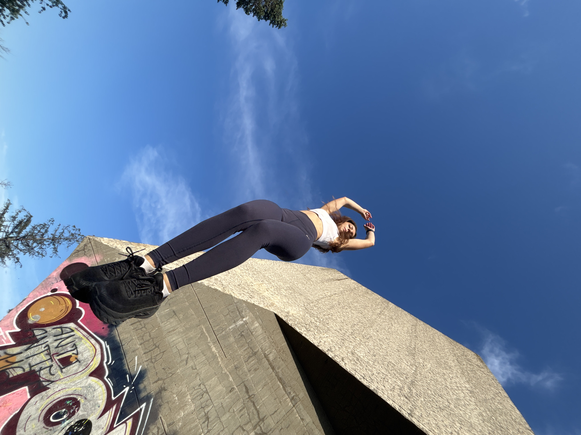 A young woman with long, brown hair is captured in a low-angle shot, standing on a concrete structure with graffiti. She wears a white sports bra and dark leggings, with black sneakers and white socks. Her arms are raised above her head, and she appears to be stretching or posing. The background is dominated by a clear blue sky with scattered wispy clouds. The concrete structure has a rough, textured surface. The graffiti on the structure includes colorful lettering and designs. The scene is set outdoors, with a hint of trees in the lower left corner.