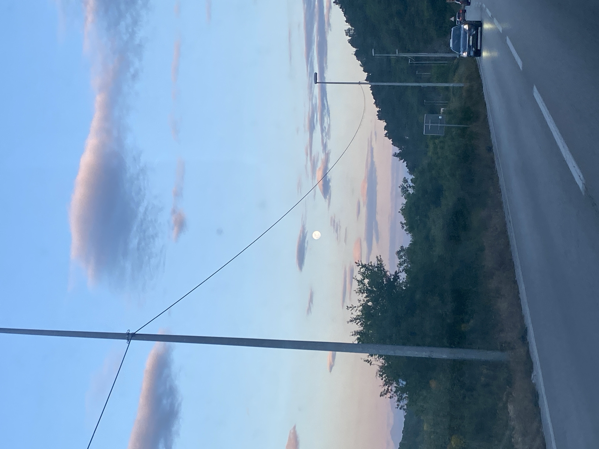 This is an outdoor photograph captured during twilight or early evening, given the sky colors and visible moon. The image is oriented vertically (or appears to be a rotated landscape shot), featuring a road on the right and a sky with scattered clouds on the left.
In the sky, shades of light blue blend with pale pink and orange, particularly towards the horizon. A distinct, full or nearly full moon is visible in the upper-left quadrant of the sky. Several elongated clouds are scattered across the expansive sky.
Running across the lower-middle of the frame is a thick, gray horizontal pole, possibly part of an electrical or communication setup. A thinner black wire is connected to this pole, extending diagonally upwards and to the left. Other thinner utility poles are visible further back on the left side of the road, one with a street light fixture attached. Behind these poles, a dark rectangular structure, possibly a sign, can be seen.
To the right, a dark asphalt road extends into the distance, marked by white dashed lines. The road surface appears somewhat reflective, suggesting dampness or the low light conditions. A dark-colored car, possibly an SUV, with its headlights illuminated, is driving on the right side of the road, moving towards the upper part of the frame.
The left side of the image features a dense expanse of dark green trees and vegetation, indicating a forested or hilly area alongside the road. No people or visible text are present in the image. The overall atmosphere is calm and transitional, capturing the moment between day and night.