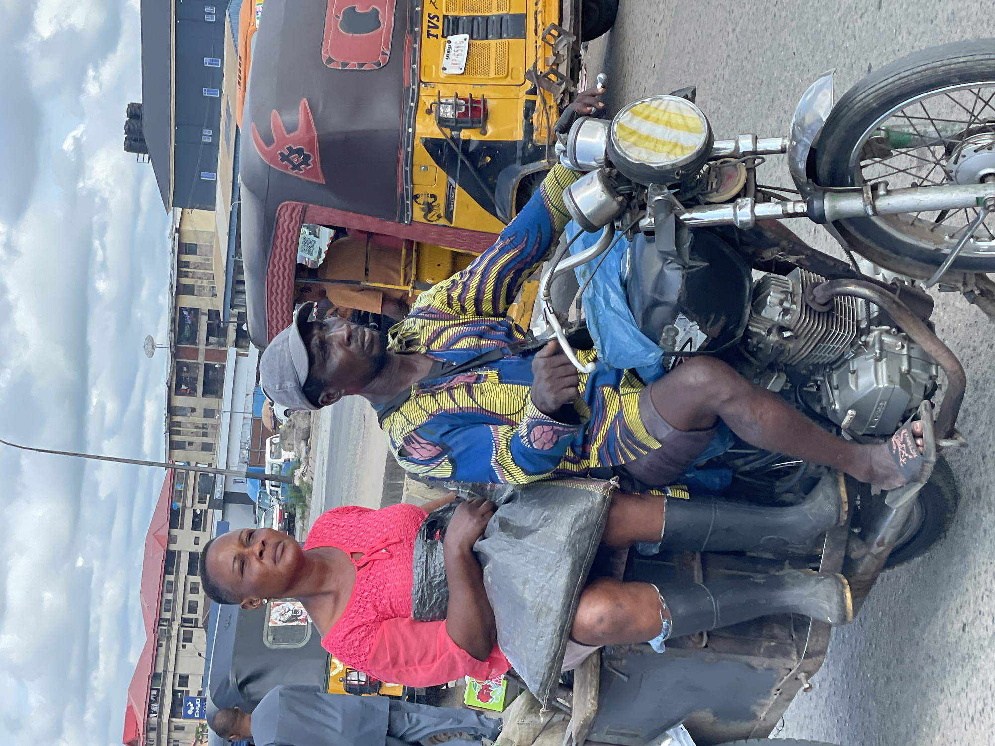 In Onitsha, Nigeria, a man wearing a colorful patterned dashiki and a grey baseball cap rides a motorcycle, with a woman in a bright pink top and black boots sitting behind him, holding a large black bag. They are in transit, with a yellow and black tuk-tuk visible behind them. The scene is set against a backdrop of urban buildings and a cloudy sky.